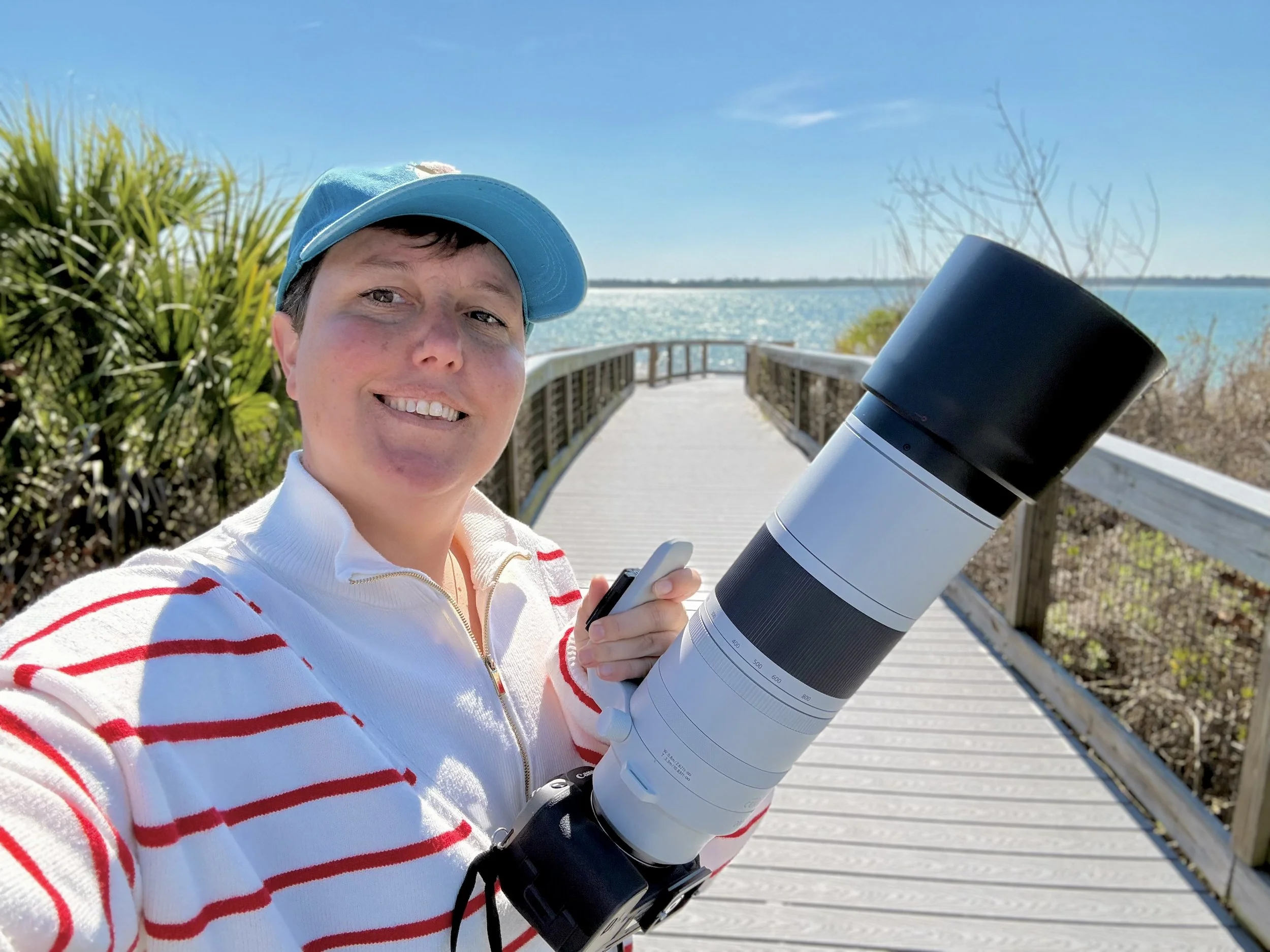 A photographer in a white sweater with red stripes and a blue cap taking a selfie with a large lens on a boardwalk near water, with clear blue sky, the ocean and some greenery in the background.