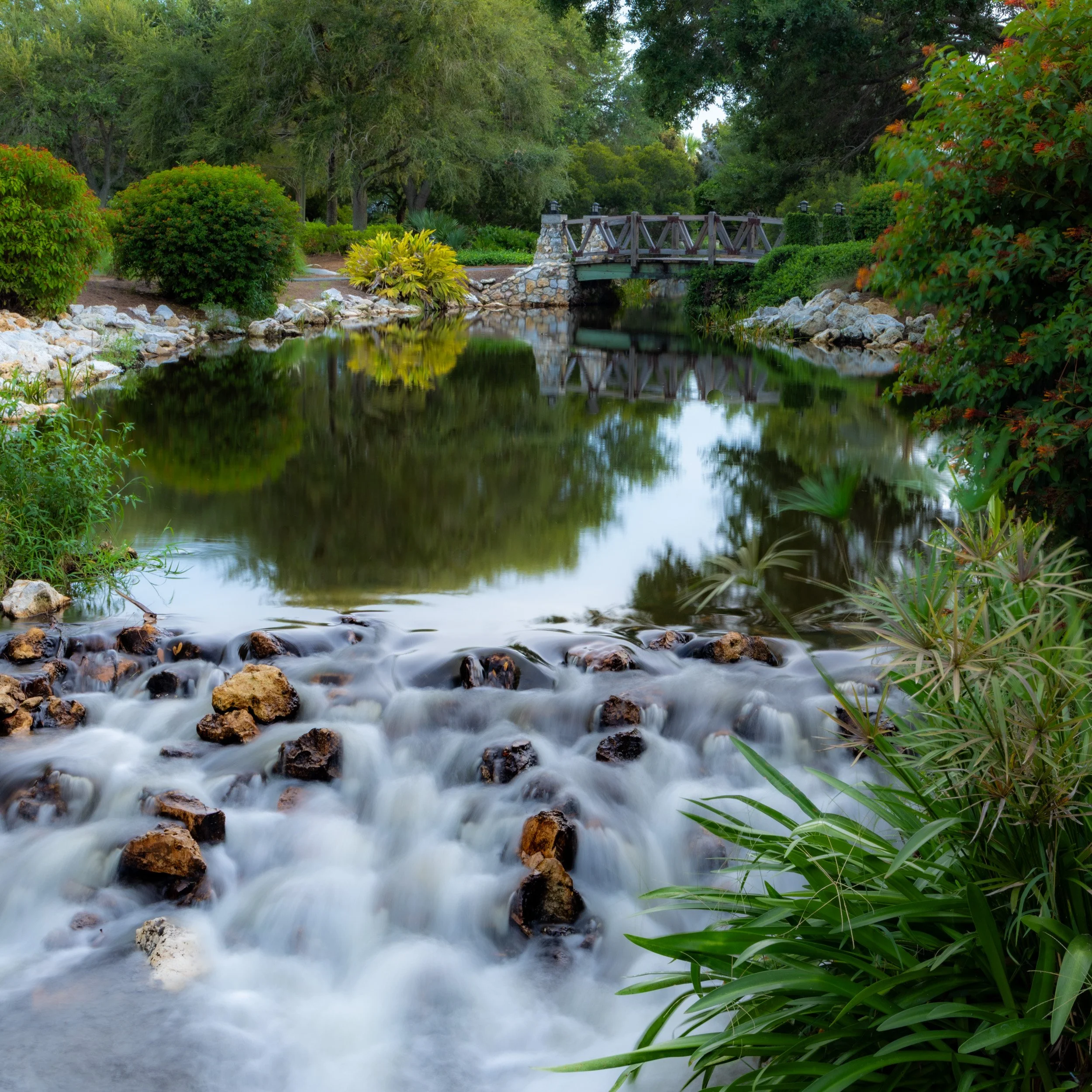 A long-exposure landscape photo of a small, rushing waterfall over rocks, leading to a calm pond that perfectly reflects a wooden footbridge and lush green trees.