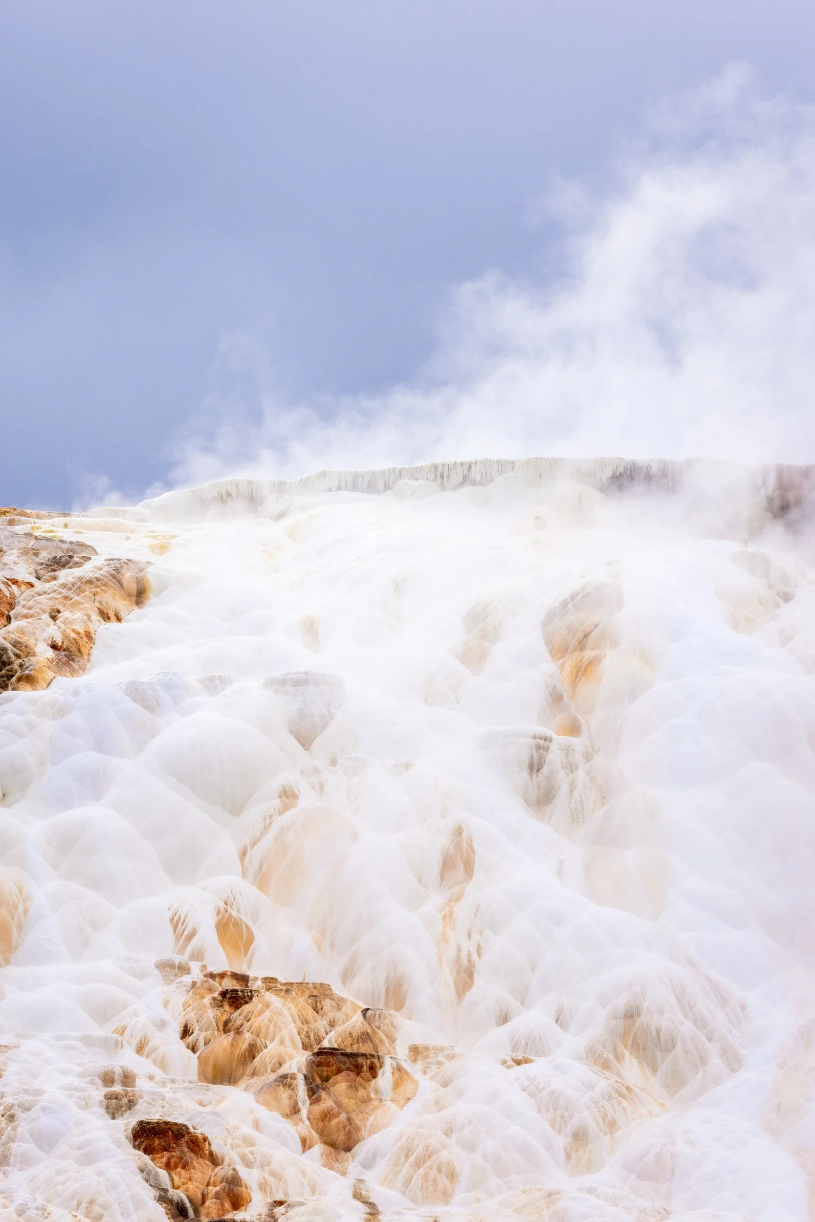 A vertical view of white mineral terraces with cascading water and thick steam rising into a dramatic sky.