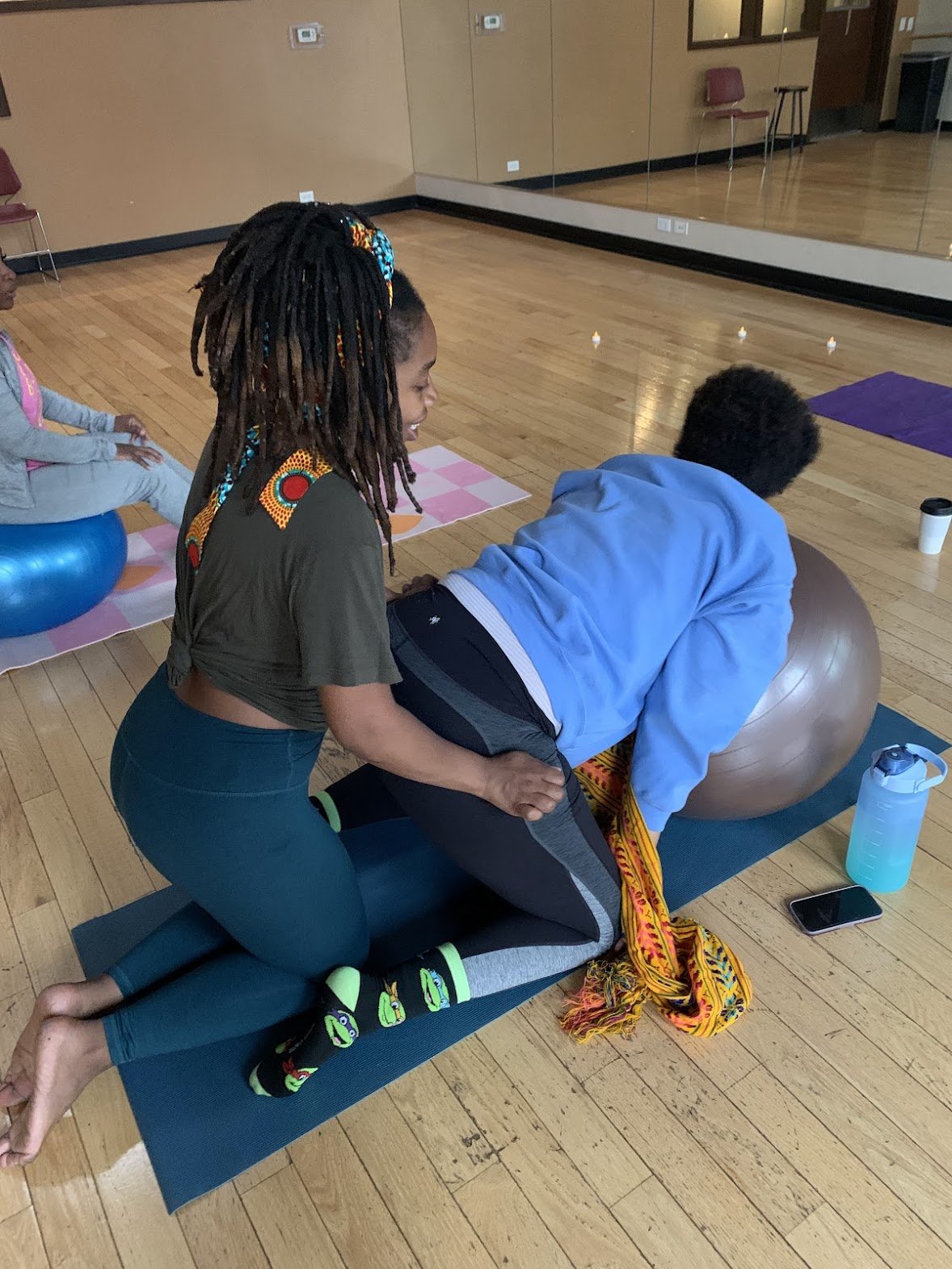 A woman assisting a man during a yoga class in a studio. The woman is kneeling beside the man, who is on a yoga mat in a child's pose, with a yoga ball nearby. In the background, there is another participant sitting on a stability ball on a different mat, and the room has a wooden floor and mirrored wall.