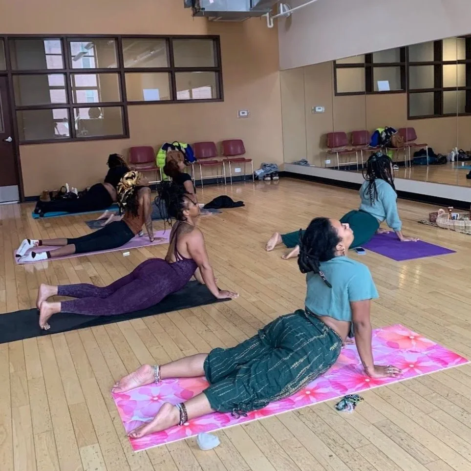 Group of women doing yoga on mats in a studio with wooden floors and a large mirror.