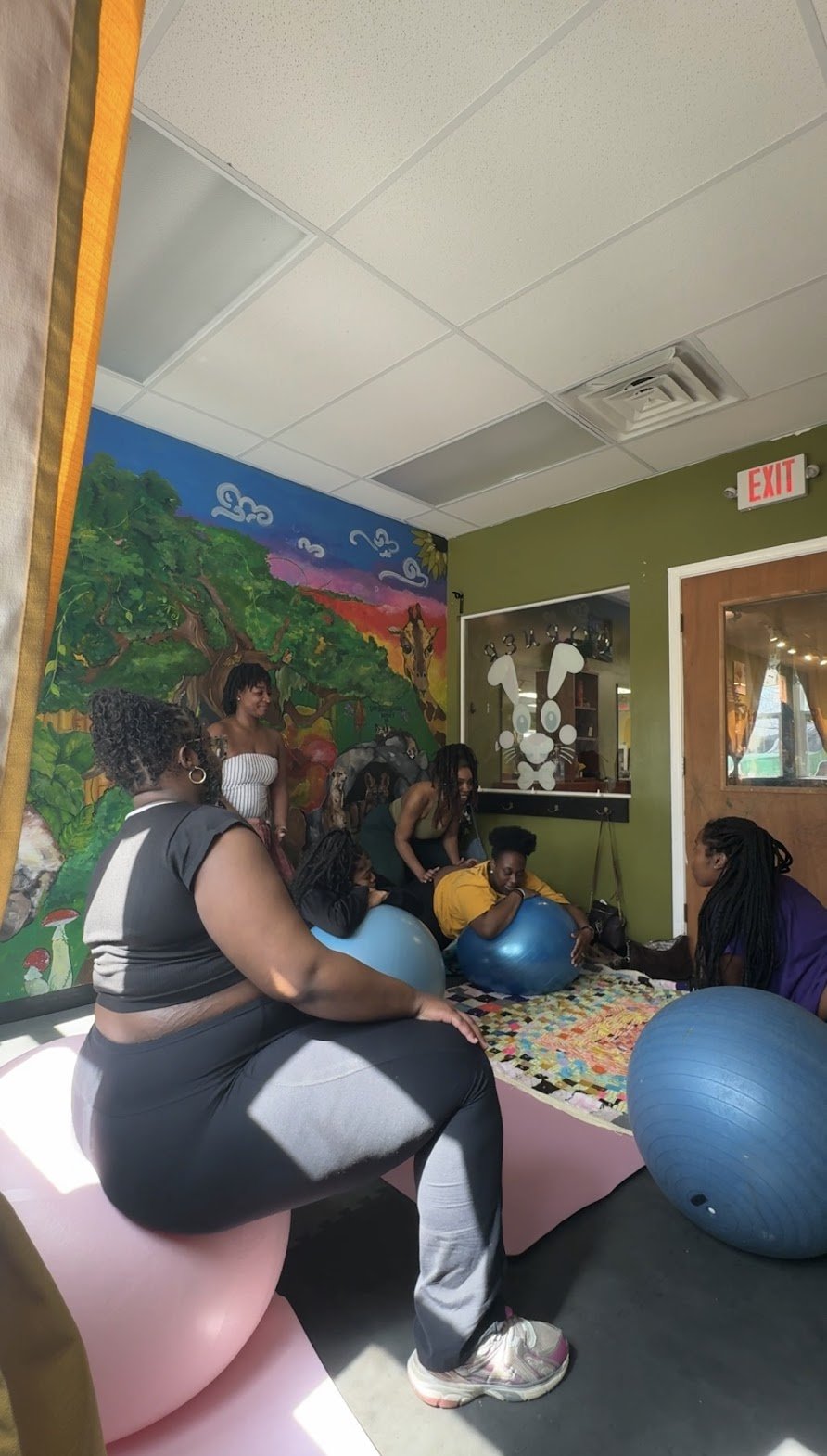 A group of women participating in a fitness class using large exercise balls in a colorful room with a mural of a forest scene on the wall.