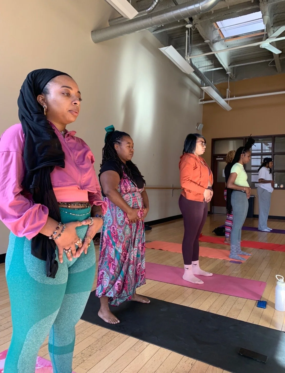 Group of women participating in a yoga class indoors, standing on yoga mats with hands in prayer position, eyes closed, focused.