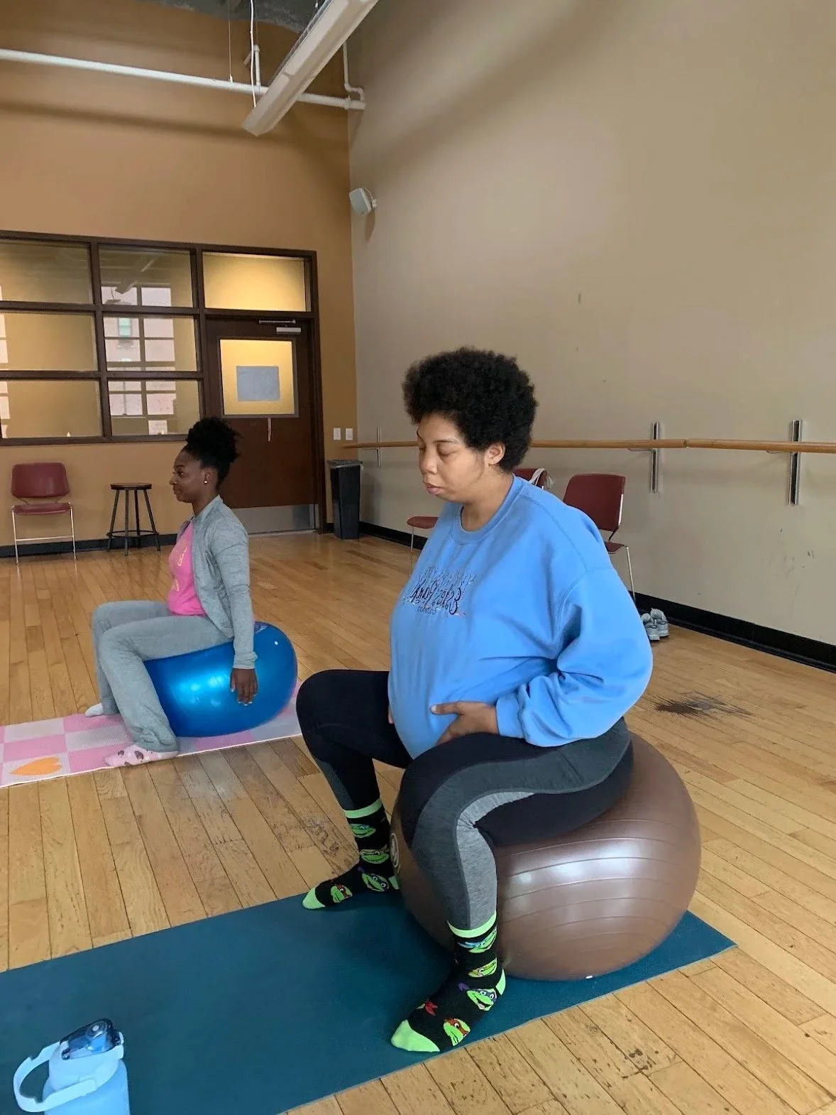 Two women in a fitness or therapy room, one sitting on a large brown exercise ball and the other on a blue exercise ball. The woman sitting on the brown ball appears to be resting or meditating, with her eyes closed. The room has a wooden floor, a ballet barre along the wall, and chairs in the background.