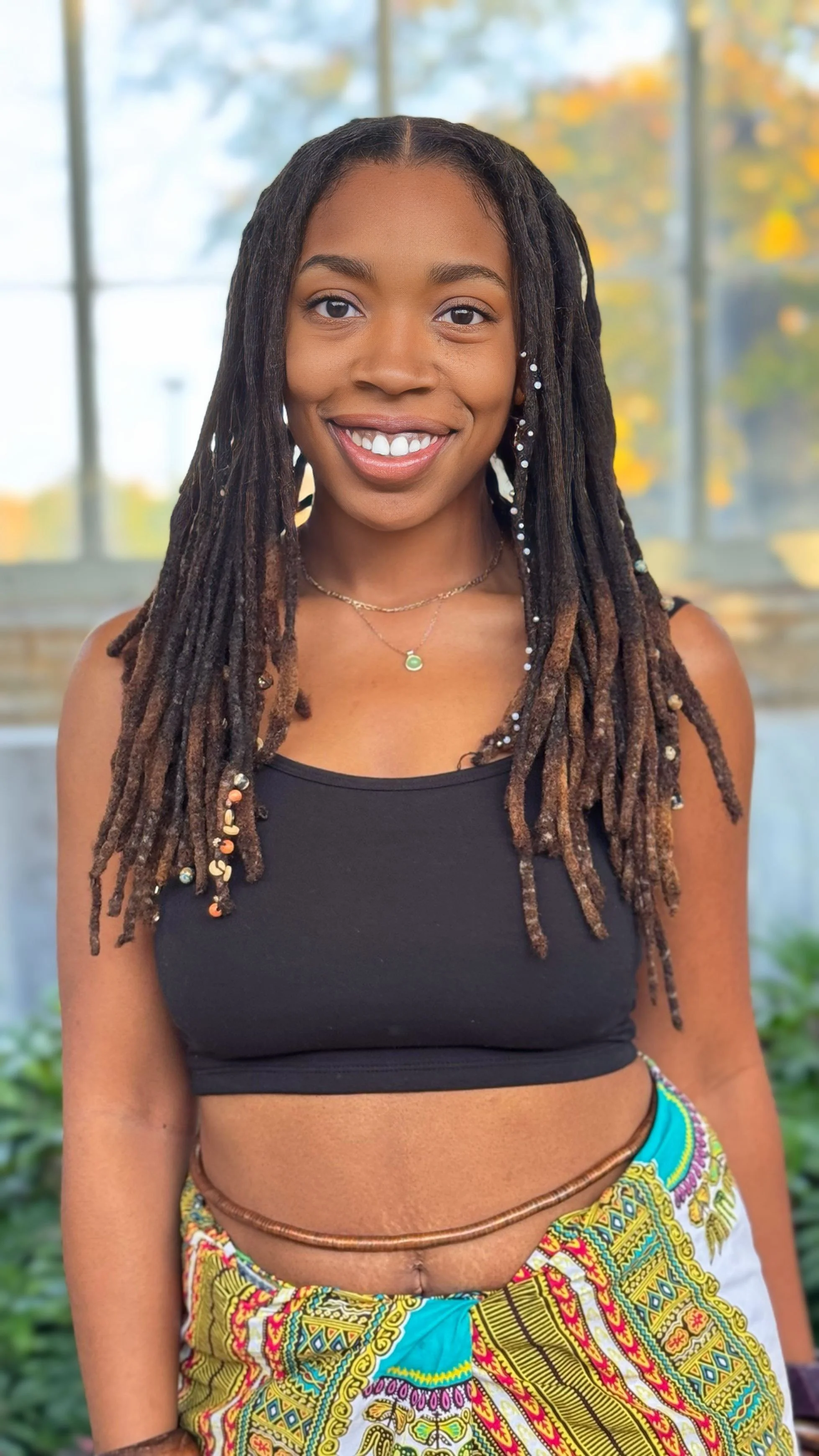 Young woman with dreadlocks smiling, wearing a black crop top and colorful skirt, standing outdoors with a background of glass windows and blurred autumn trees.