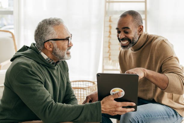 Two men, one older with gray hair and glasses, and one younger, smiling and leaning towards a tablet, in a bright room with natural light. advocacy, support, care