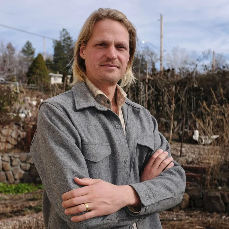 A man with shoulder-length blond hair and a light beard, wearing a gray jacket over a plaid shirt, standing outdoors with crossed arms, in front of a garden or backyard with trees and fencing.