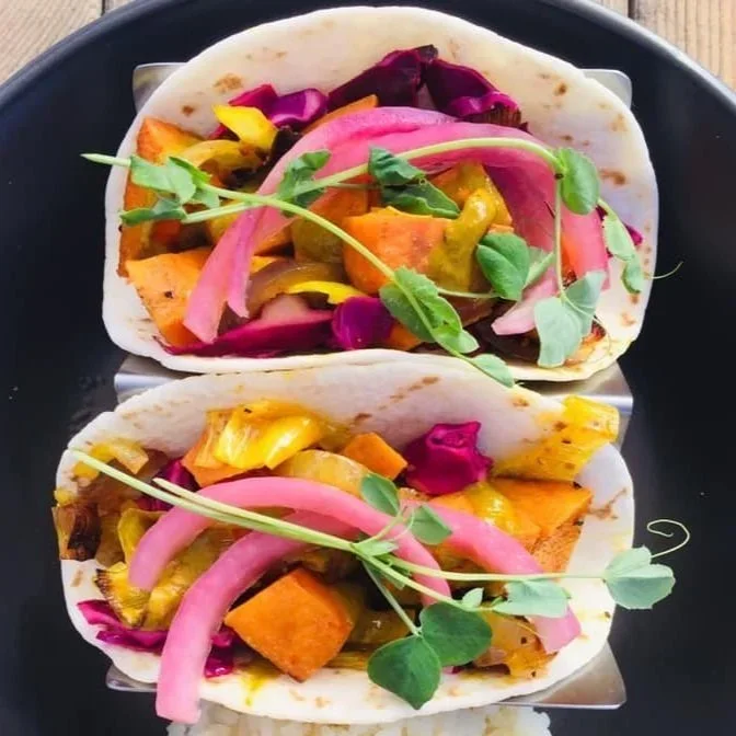 Two tacos filled with roasted vegetables, pickled onions, and topped with microgreens on a black plate.