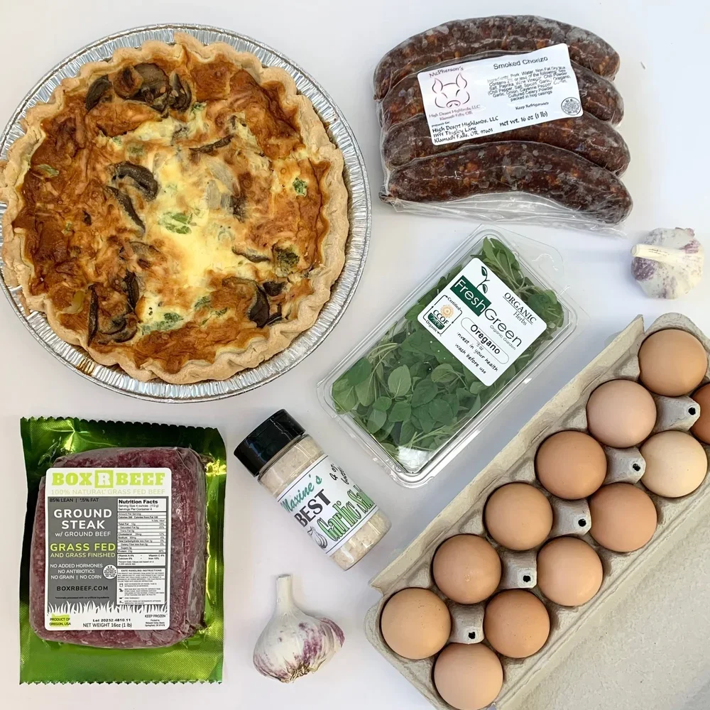 A pie with mushrooms and greens on a white surface, a package of smoked chorizo sausages, a container of fresh oregano, a carton of farm-fresh eggs, a bag of ground beef, a head of garlic, and a container of garlic powder.