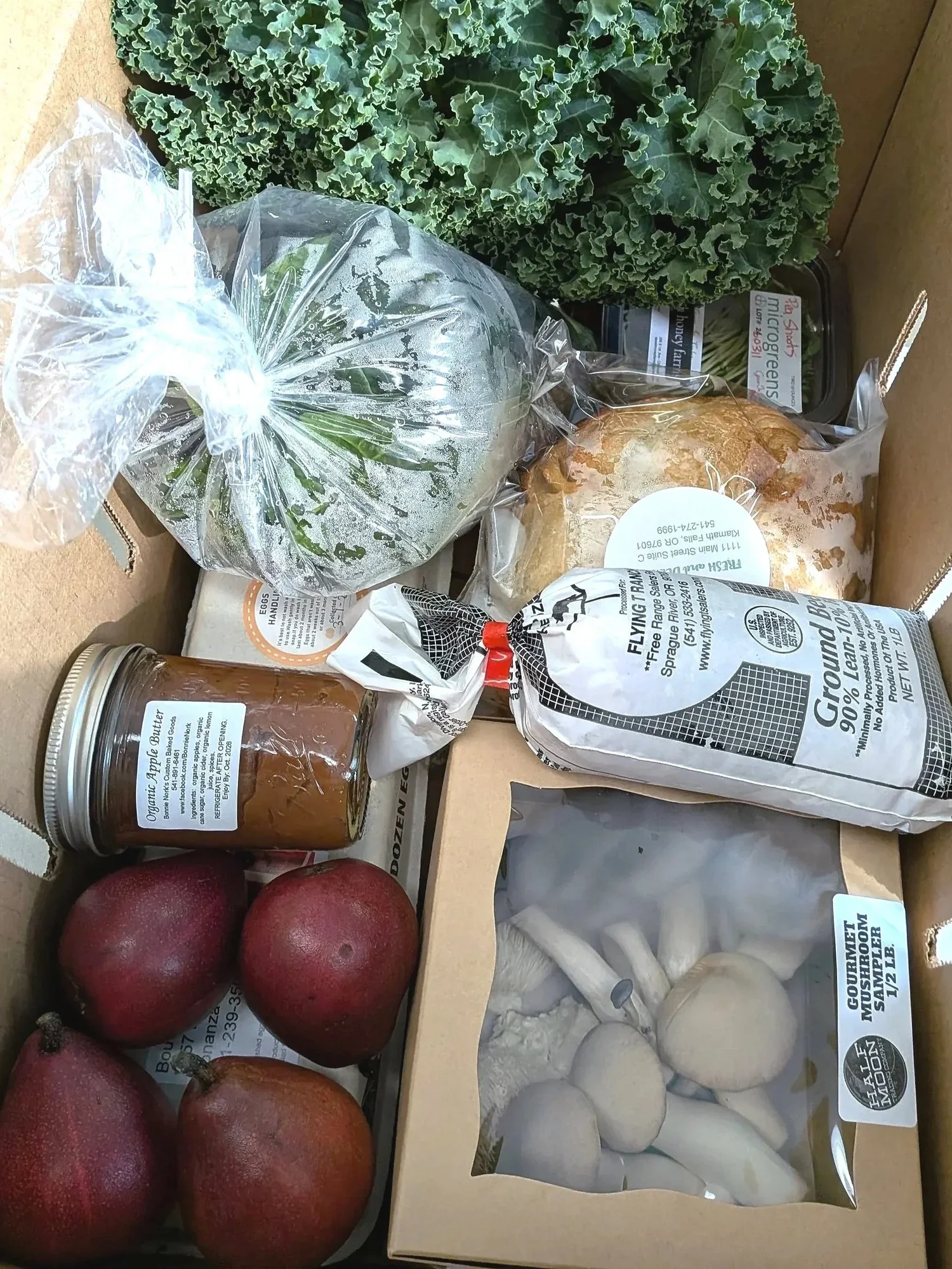 Green leafy kale, a bag of fresh herbs, a loaf of baked bread, a jar of organic apple butter, red apples, a box of white mushrooms, a bag of ground beef, a bundle of scallions, and a package of eggs.