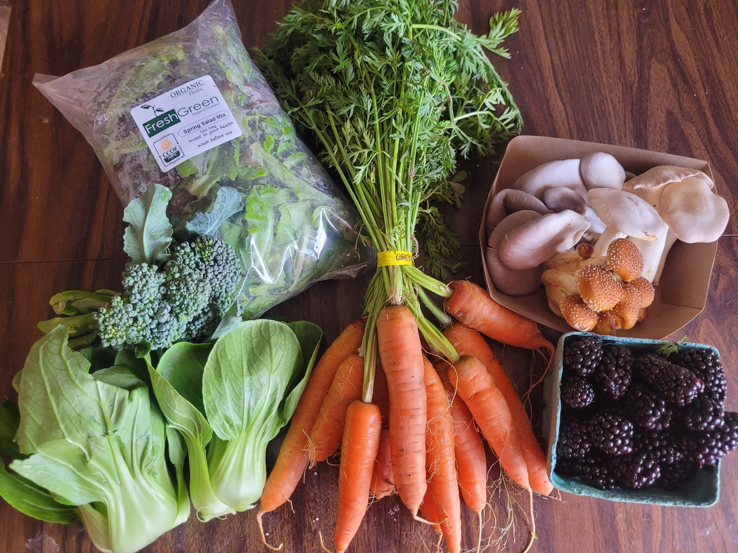 Fresh garden vegetables including carrots, broccoli, baby bok choy, mixed greens, mushrooms, blackberries, and shimeji mushrooms on a wooden surface.