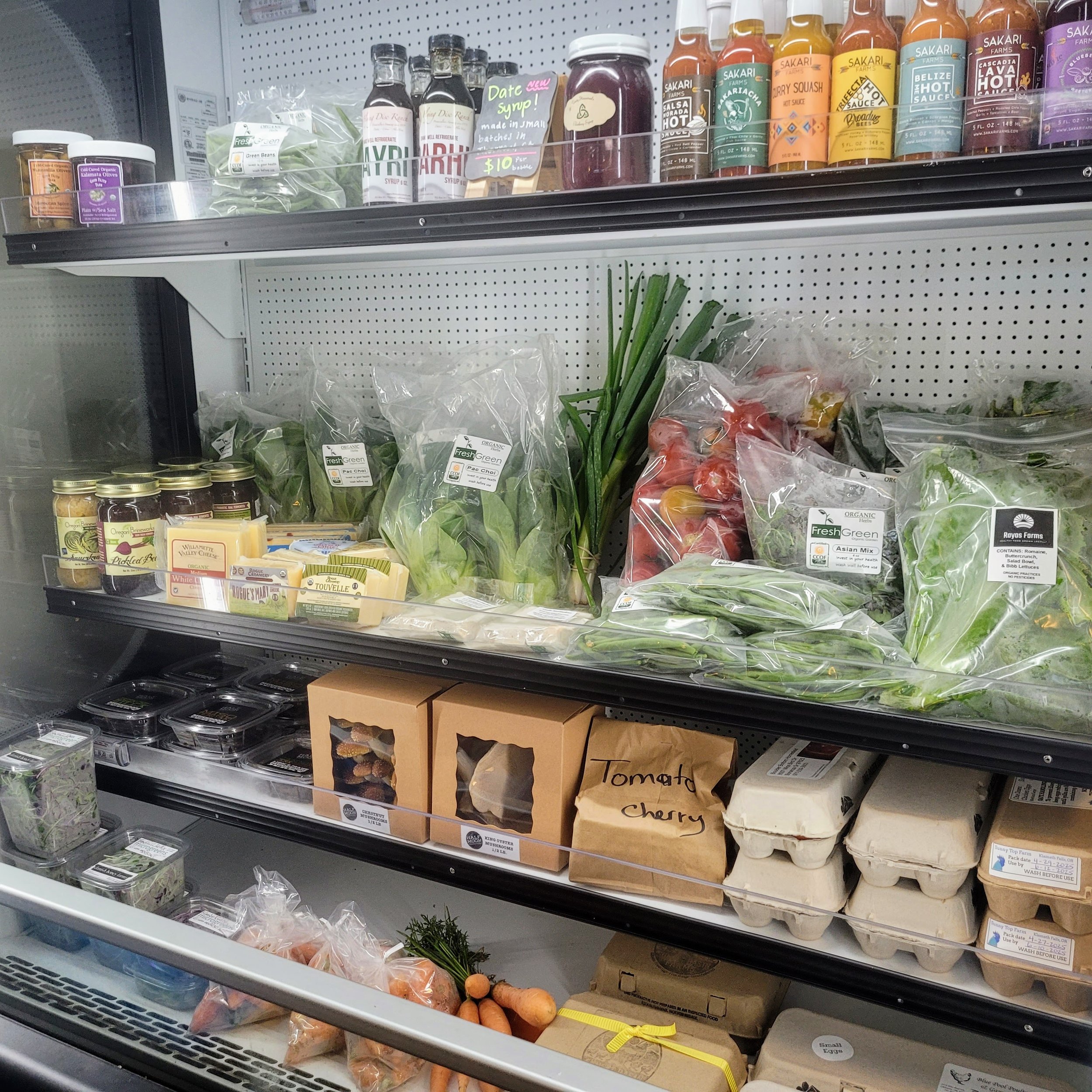 Refrigerated produce section with leafy greens, cherry tomatoes, eggs, and packaged vegetables, and shelves with bottles of sauces and jams.