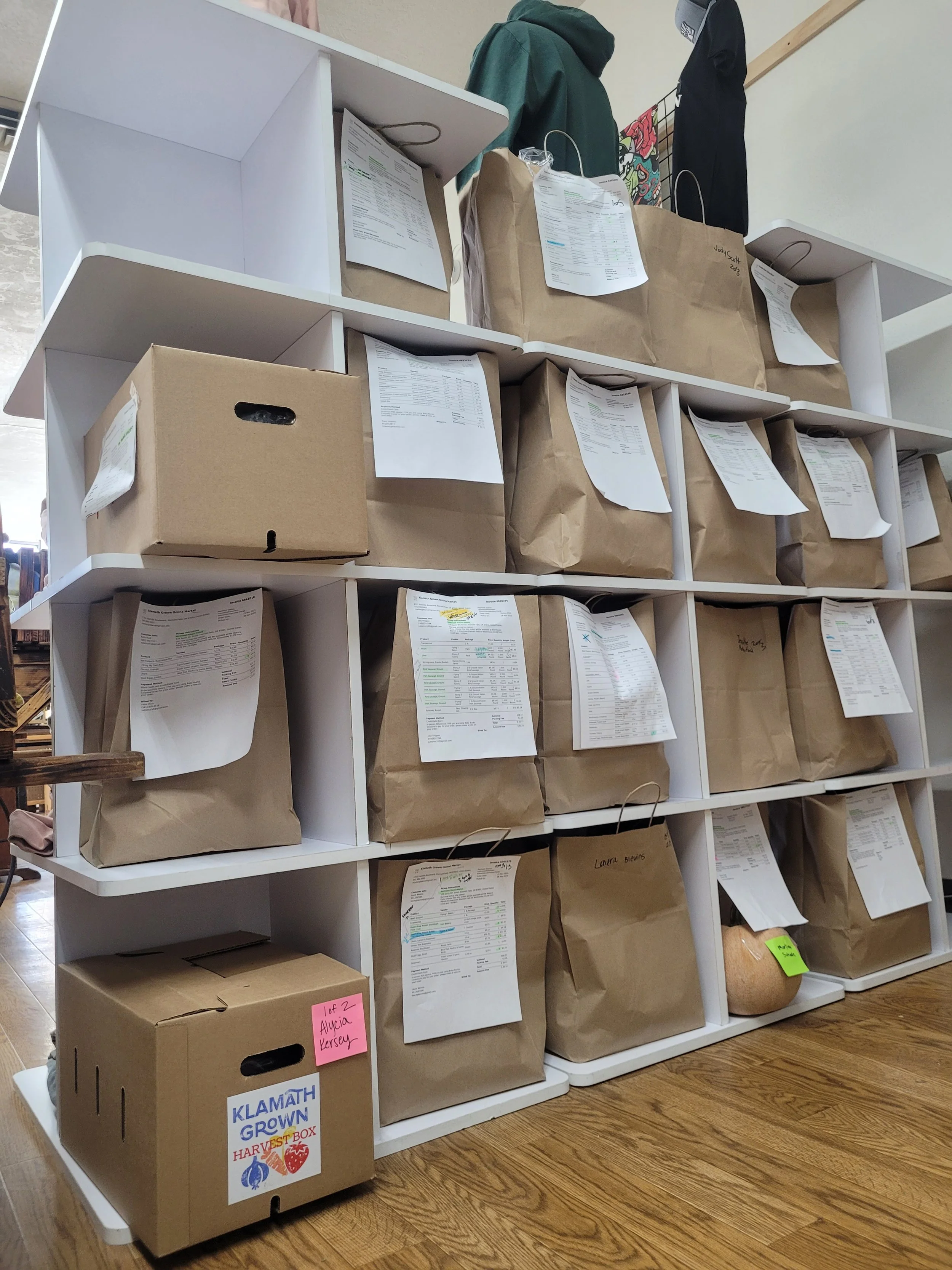 Shelving unit filled with brown paper bags and a few cardboard boxes, paper receipts attached to the bags, with clothing items hanging on the top shelf.