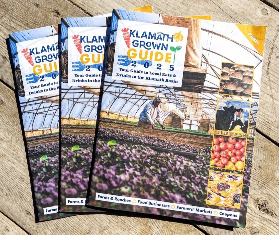 Three copies of the Klamath Grown Guide 2025, showing a greenhouse with a farmer planting crops, along with small images of apples, cattle, and baked goods, on a wooden surface.