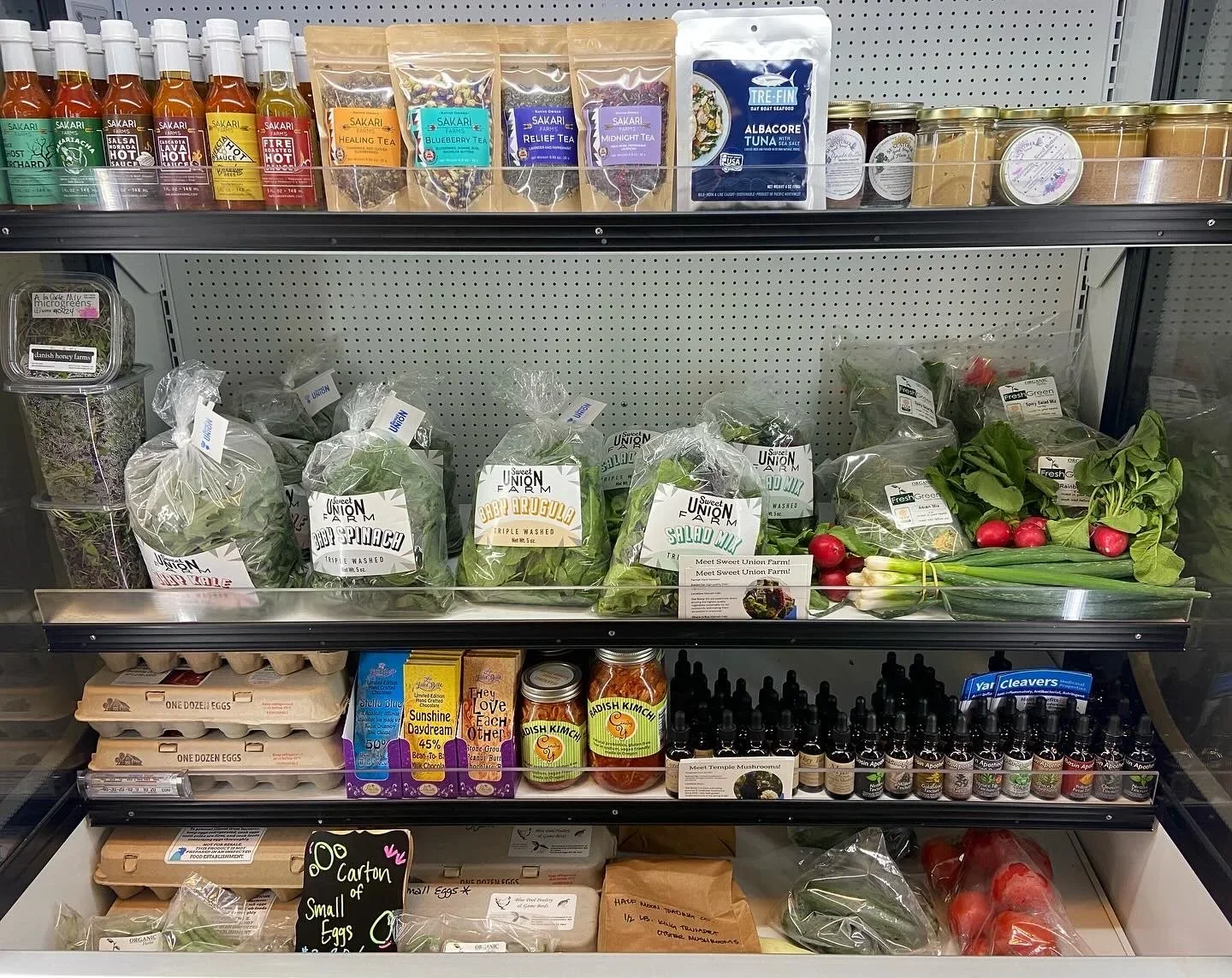 Display of fresh produce, such as radishes, spinach, arugula, lettuce, and leeks, along with eggs, herbs, and various bottled products on supermarket shelves.