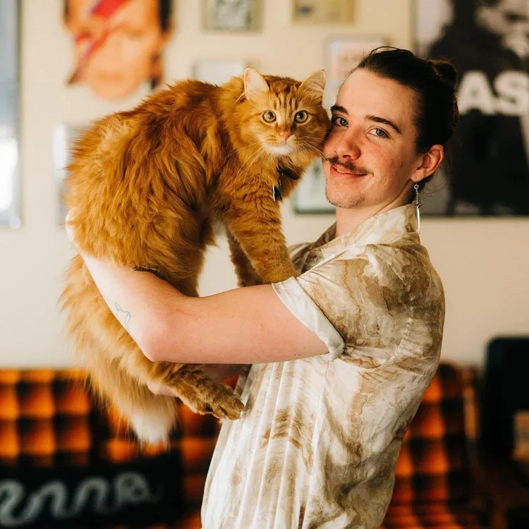 A man with dark hair and a mustache holding a large orange Maine Coon cat indoors, with blurred artwork on the wall in the background.
