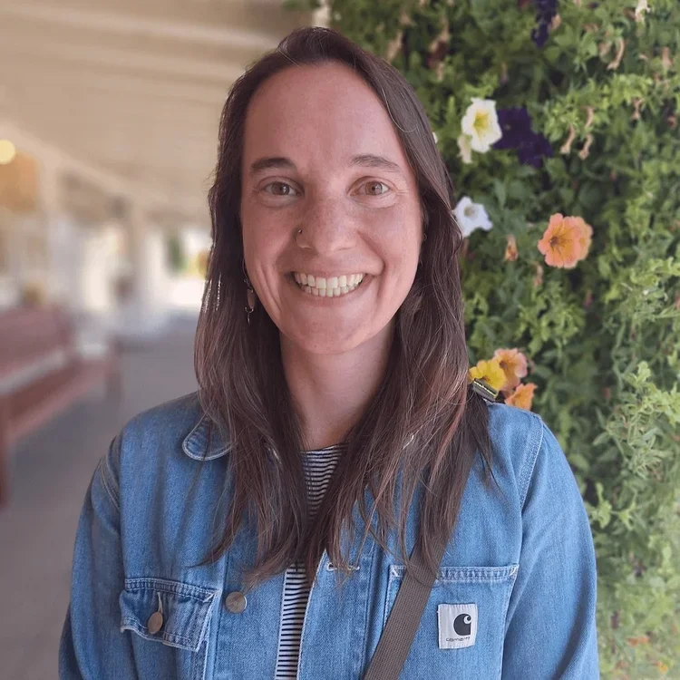 A smiling woman with long brown hair, wearing a denim jacket, standing in front of a green plant with colorful flowers.