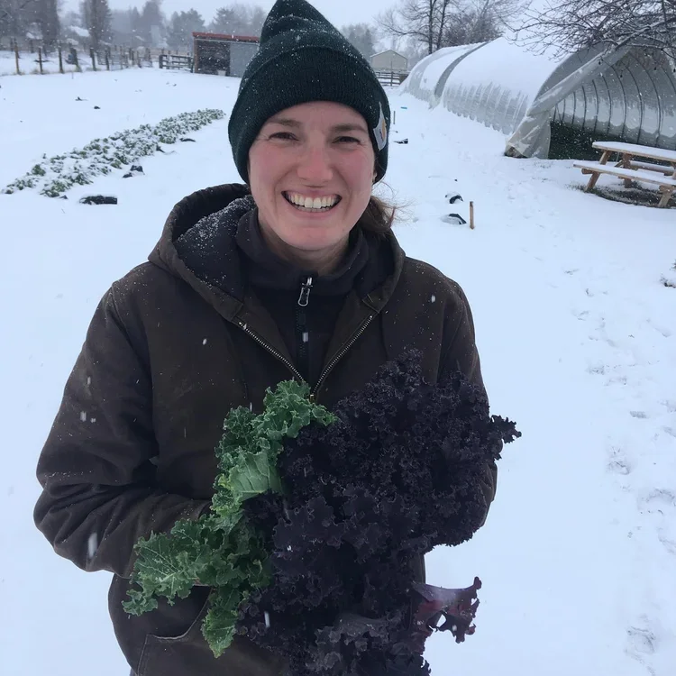 A woman smiling outdoors in snowy weather, wearing a black beanie and brown jacket, holding fresh kale and purple kale.