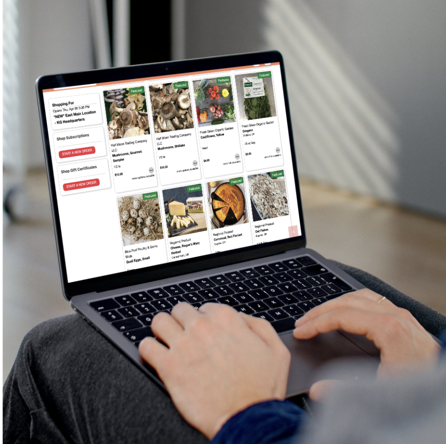 Laptop screen displaying an online grocery shopping website with various food items including mushrooms, strawberries, greens, eggs, cheese, a pie, and bread.