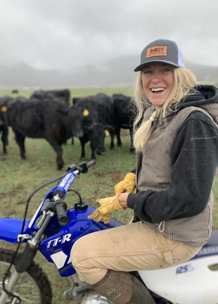 A woman in outdoor attire sits on a blue dirt bike on a farm, smiling at the camera, with a herd of black cows in the background under a cloudy sky.