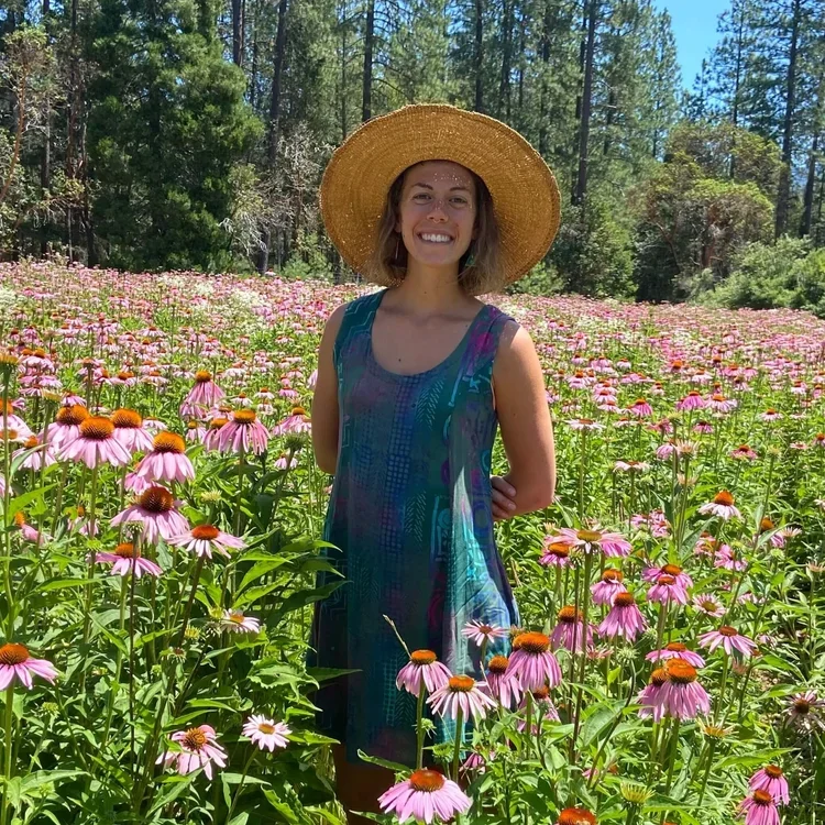 A young woman in a blue patterned summer dress and a wide-brimmed straw hat standing in a field of pink coneflowers, smiling at the camera with trees and a bright blue sky in the background.