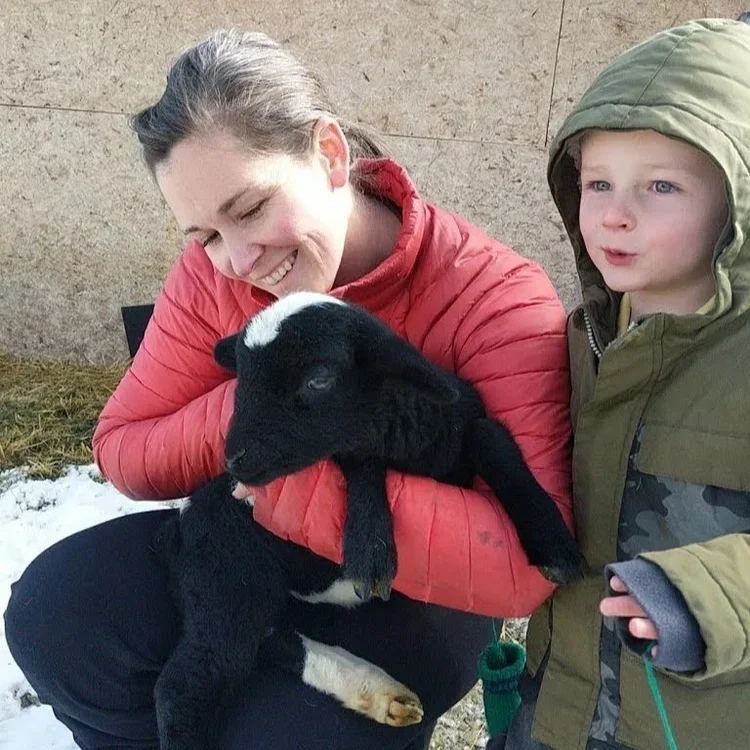 A woman in a red jacket smiling and holding a black and white baby goat, with a young boy in a green jacket standing next to her outside on a snowy ground.
