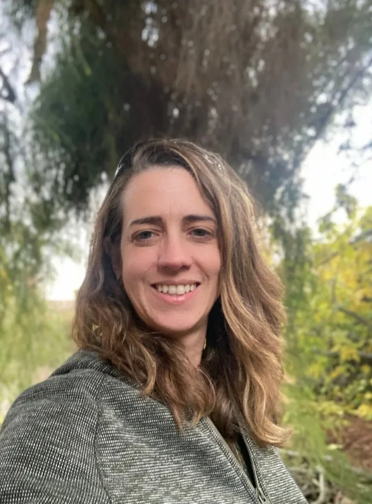 A woman with wavy brown hair smiling outdoors in front of trees and greenery.