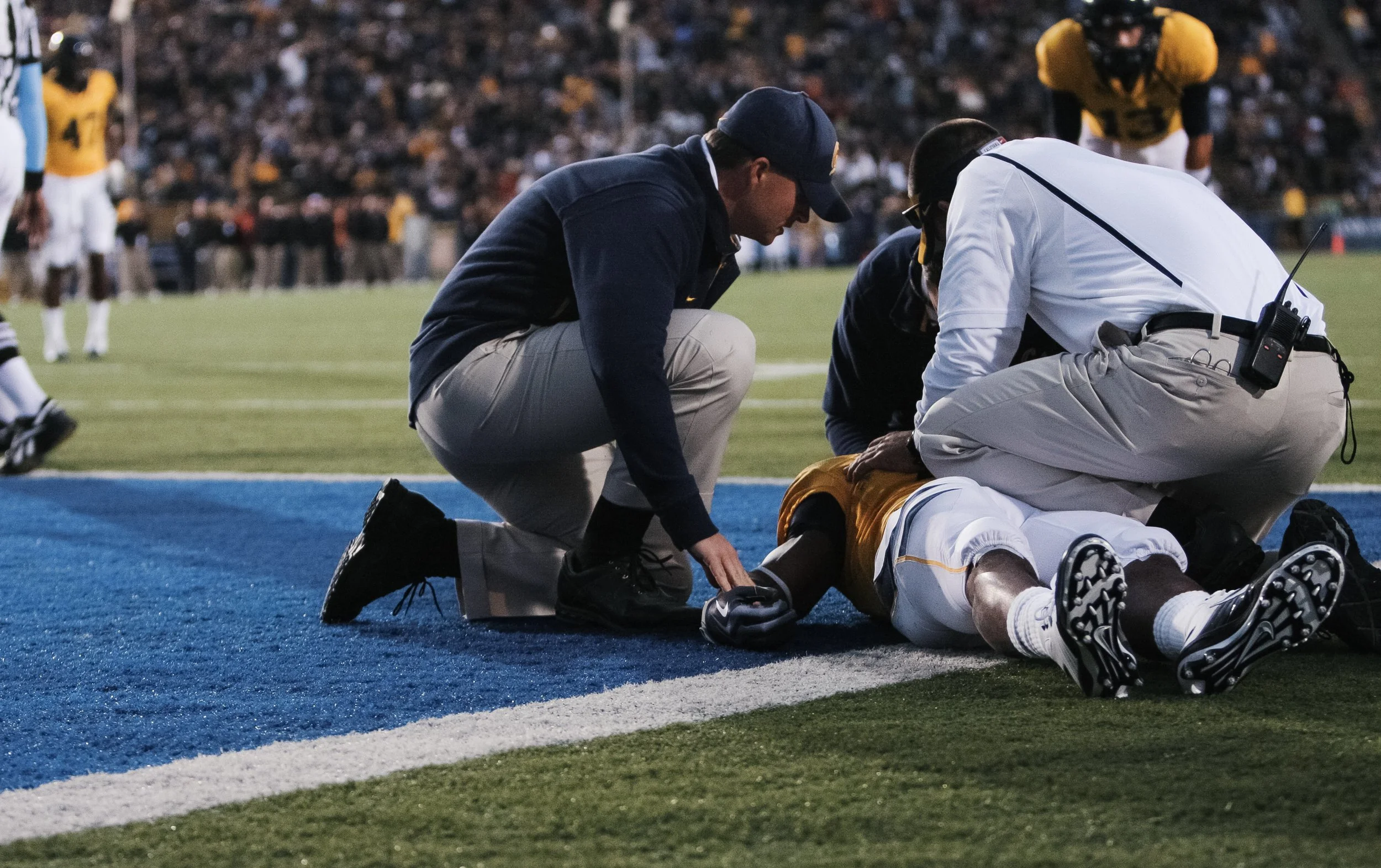 Football player lying on the ground being attended by medical staff during a game, with another player in yellow jersey nearby and a crowd in the background.