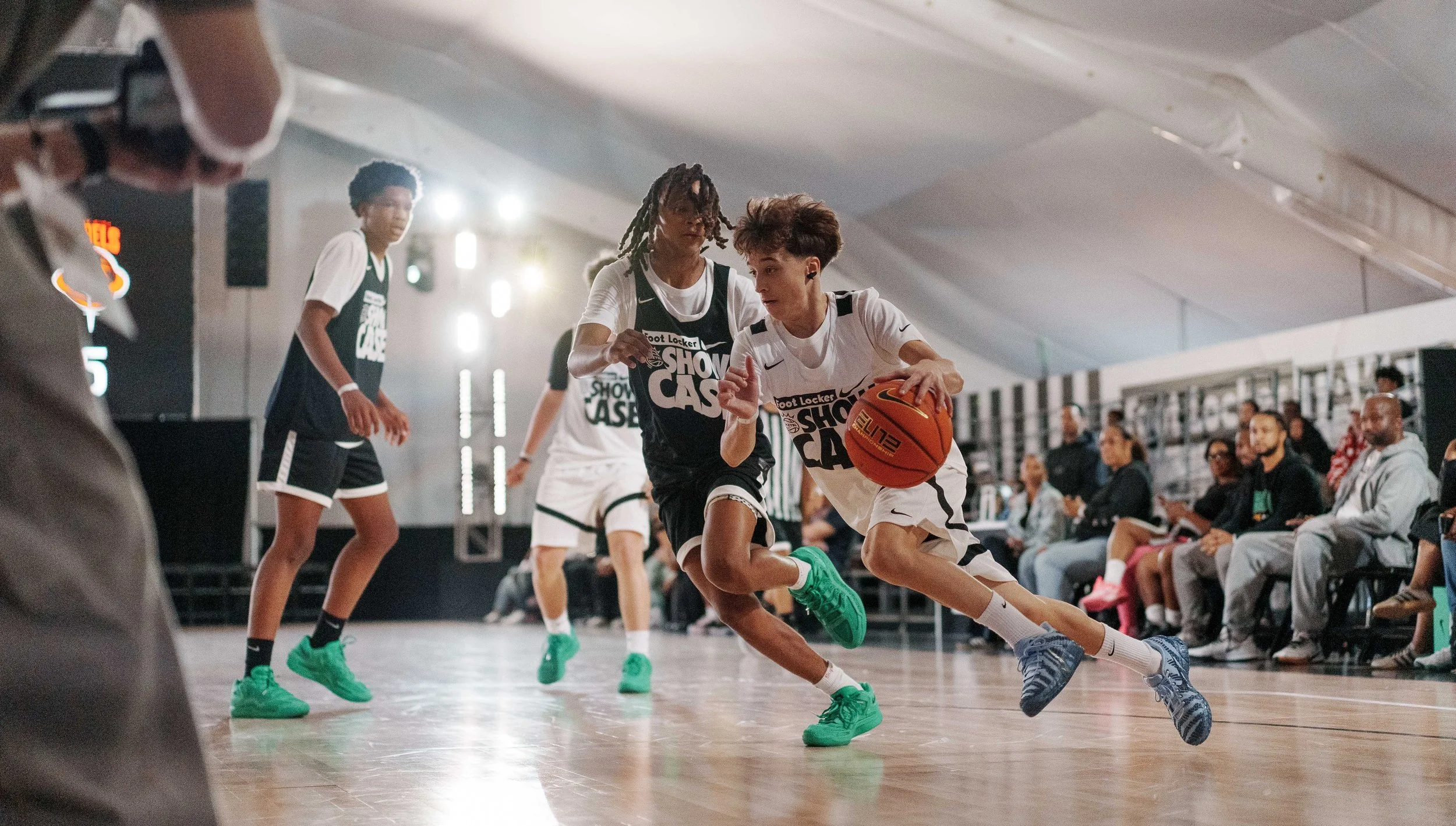 Two young male basketball players in white jerseys and green shoes compete for the ball during a game, with an audience seated in the background.