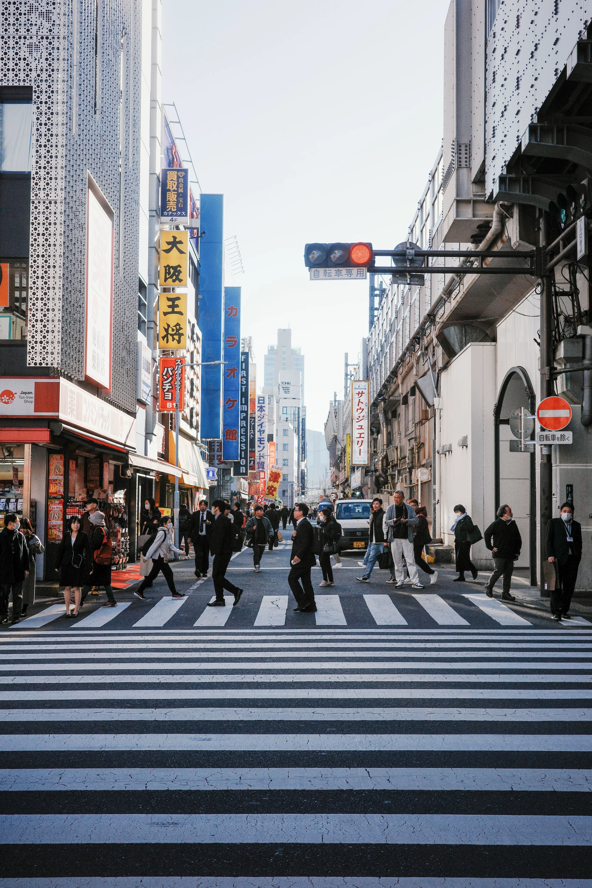 A busy city street crossing with pedestrians walking on a zebra crosswalk, surrounded by tall buildings with colorful signage in Japanese and English, and a traffic light showing red.