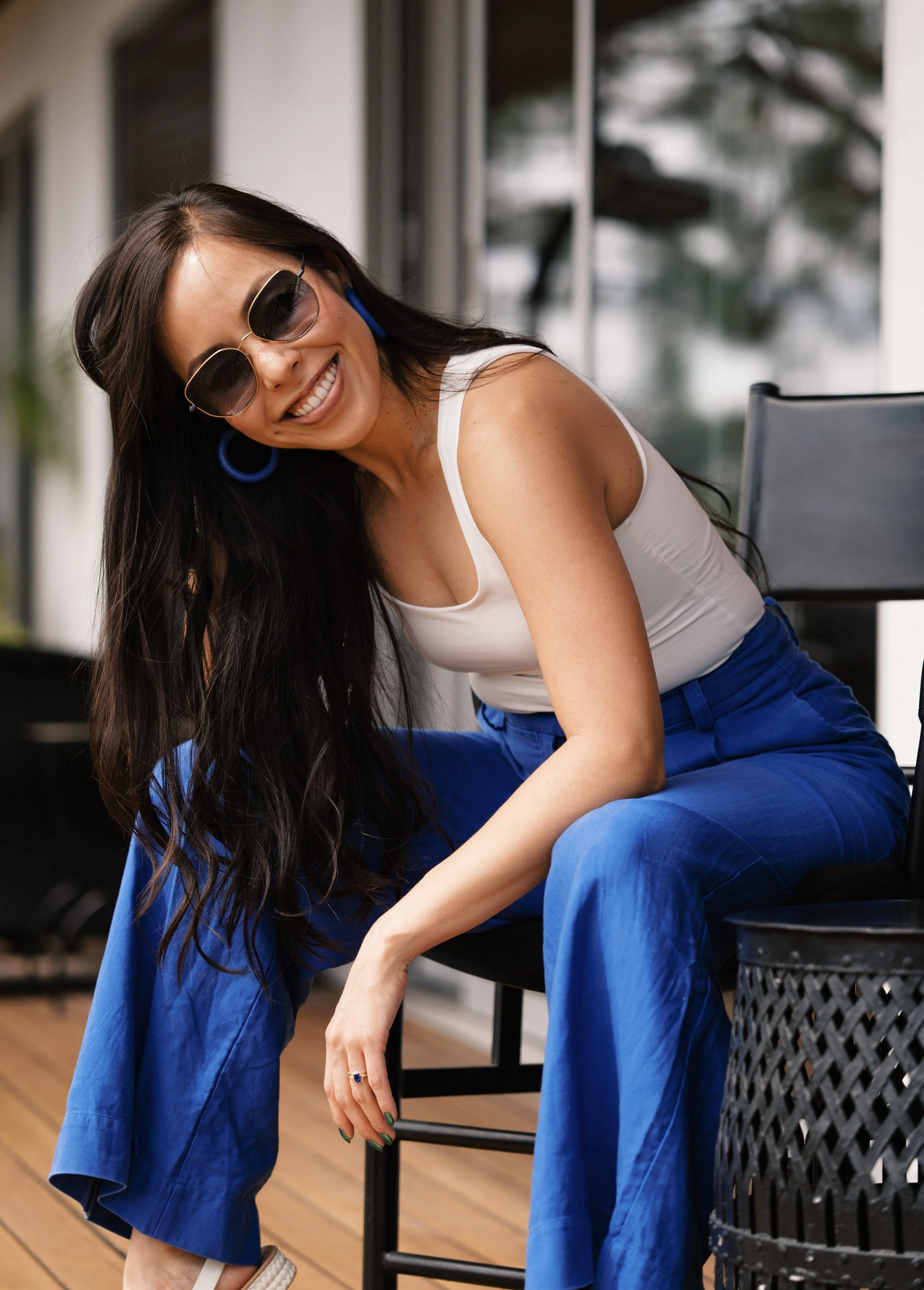 A woman smiling and wearing sunglasses, a white tank top, and blue pants, sitting on a black chair in a well-lit room with large window and wooden floor.