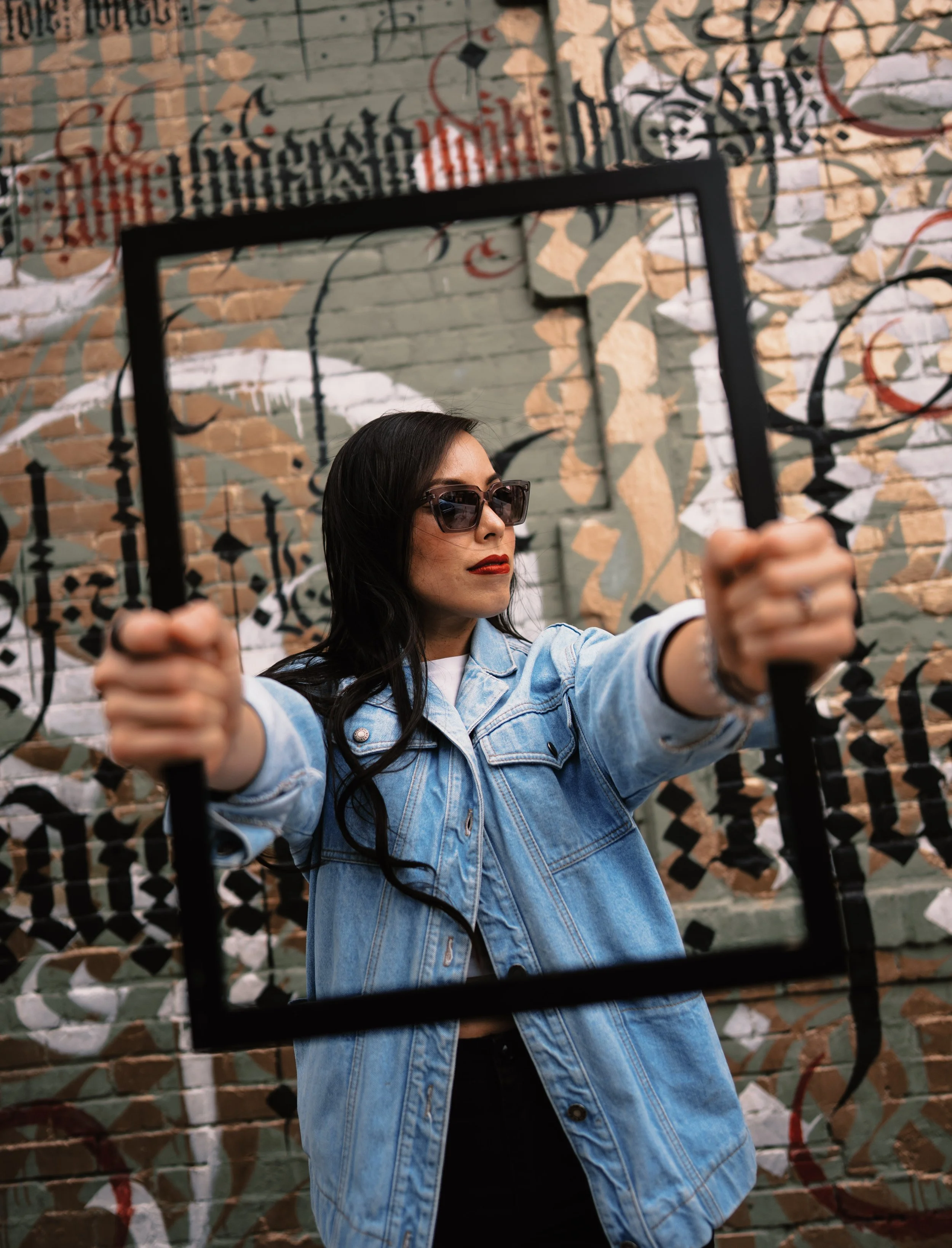A woman with dark hair and sunglasses taking a selfie in front of a graffiti-covered brick wall, holding a black frame in front of her.