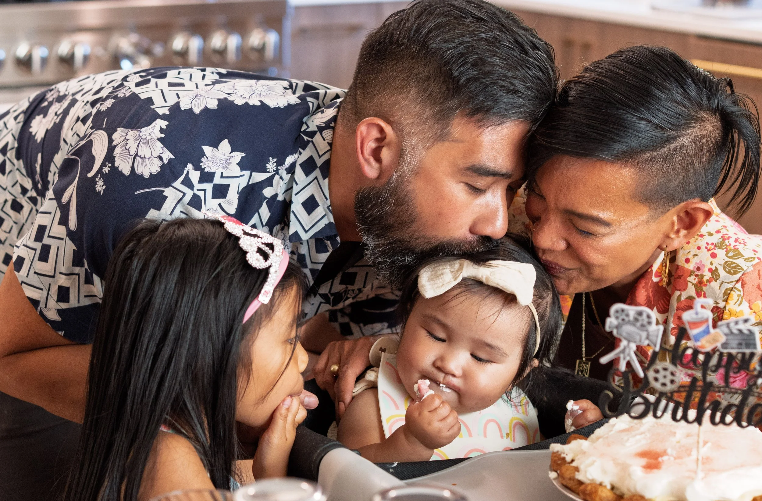 A man, woman, and children are gathered around a table, sharing a moment while cutting a birthday cake together. The children are eating slices of cake, and everyone appears happy and engaged in a celebration.