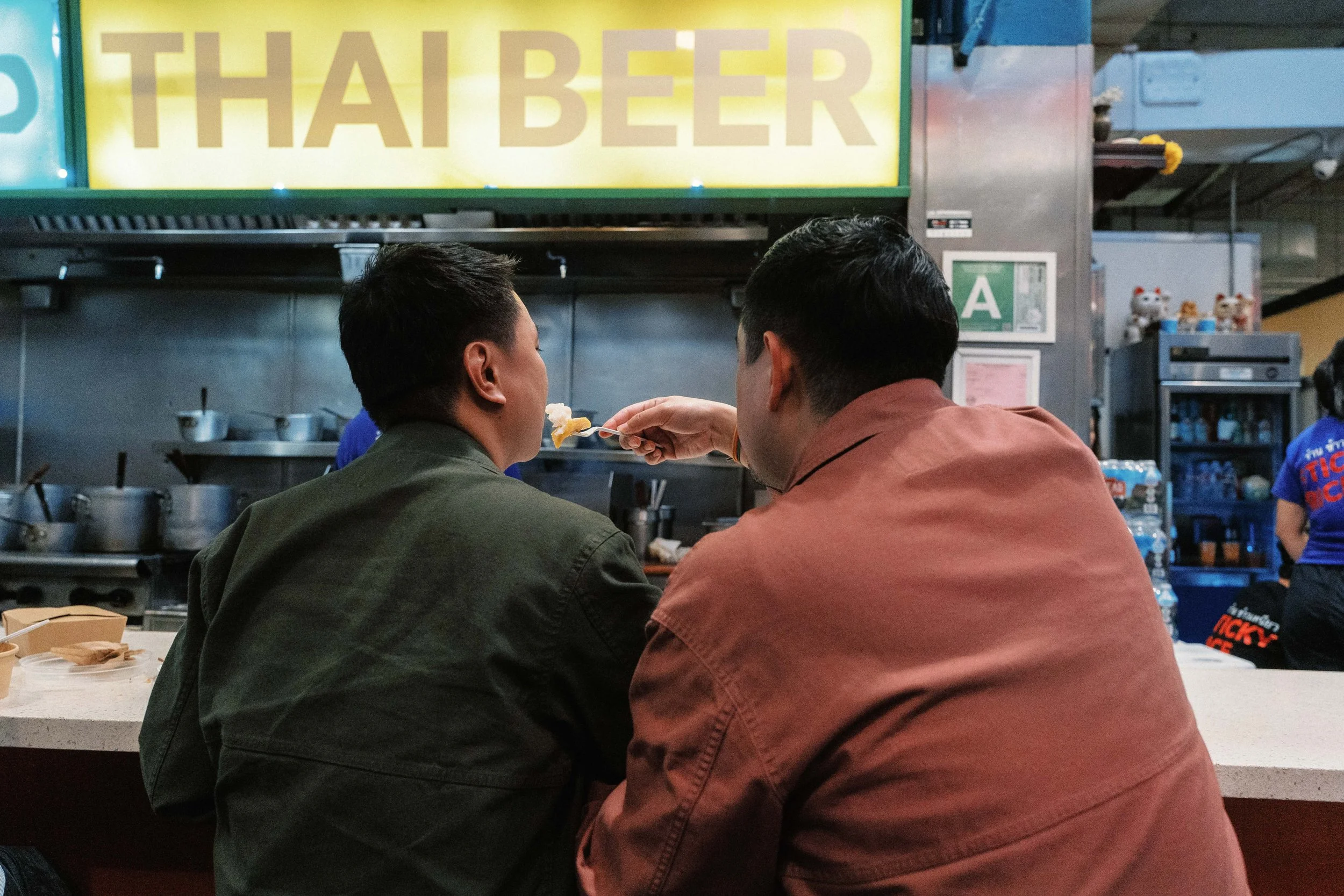 Two people sitting at a counter in a restaurant or cafe, with one person feeding the other. Behind them, a yellow sign reads 'TAH BEE' and kitchen utensils are visible.