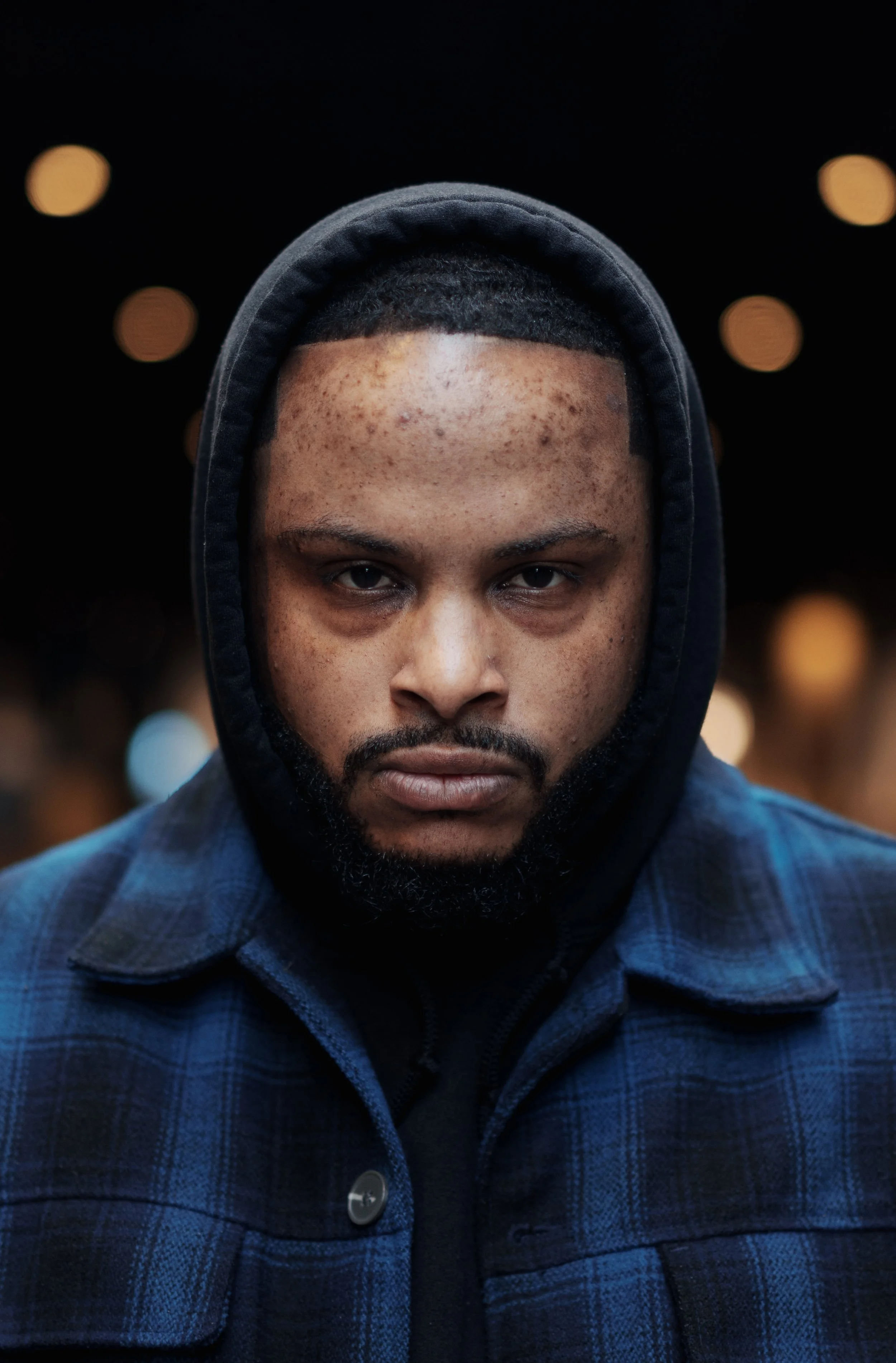 Close-up of a man wearing a hoodie and a plaid blue jacket, looking directly at the camera with a serious expression. The background is black with blurred circular lights.