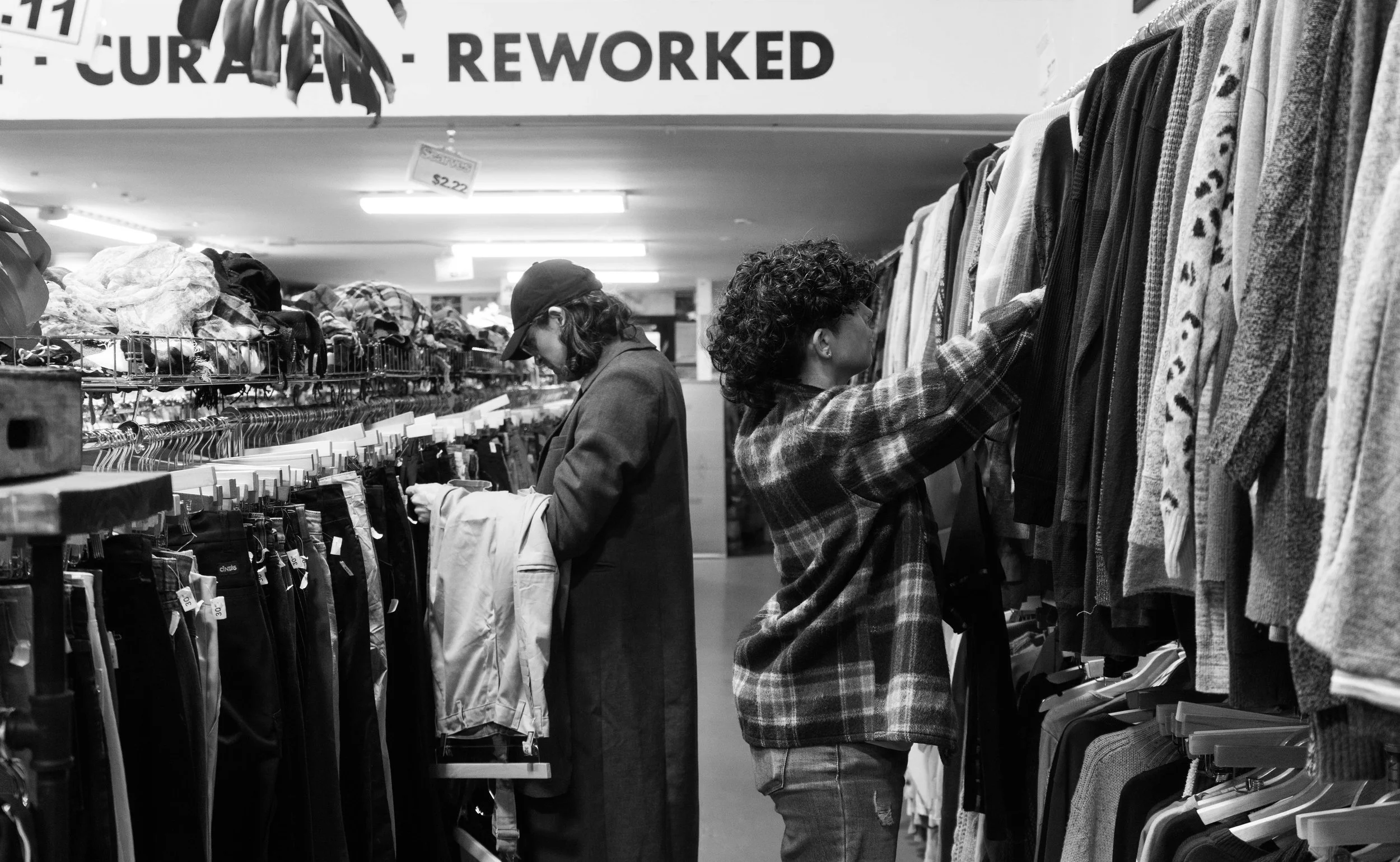 Two women shopping for clothes in a thrift store or clothing store aisle, looking at shirts on a clothing rack.