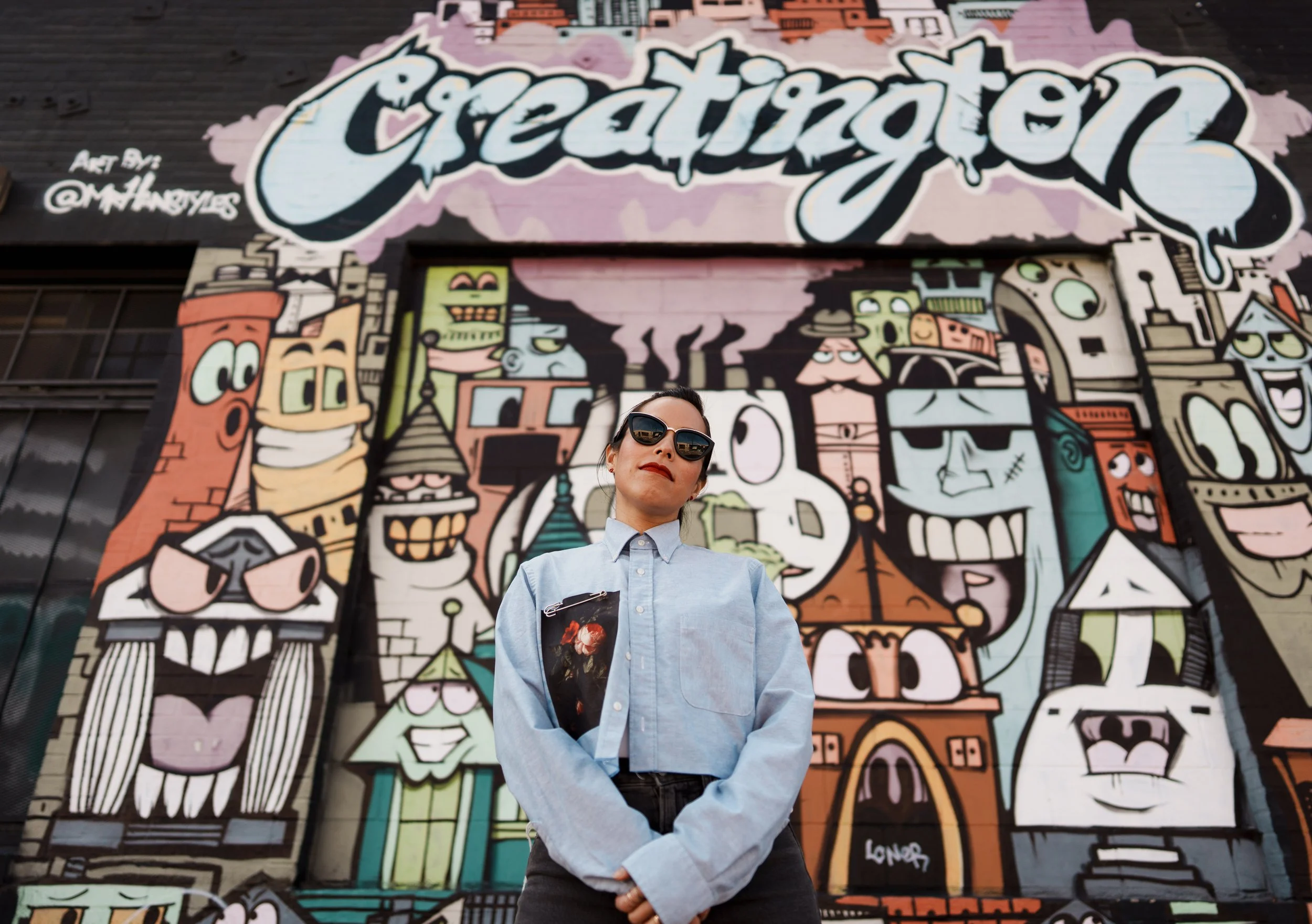 A woman wearing sunglasses and a light blue shirt standing in front of a colorful graffiti mural that reads 'Creat Ignition' with stylized cartoon characters and cityscape elements.
