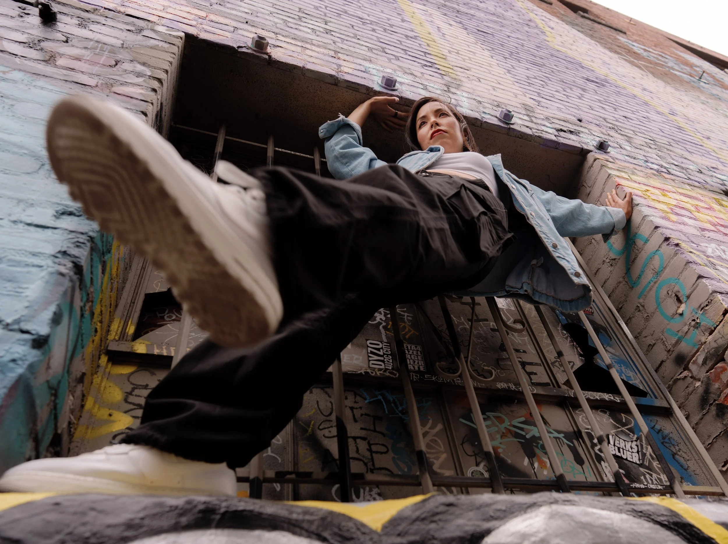 A person lying on their back on a graffiti-covered rooftop, looking up with one arm behind their head and the other resting on the edge of the roof. They are wearing a denim jacket, a white t-shirt, and dark pants, with sneakers visible at the bottom