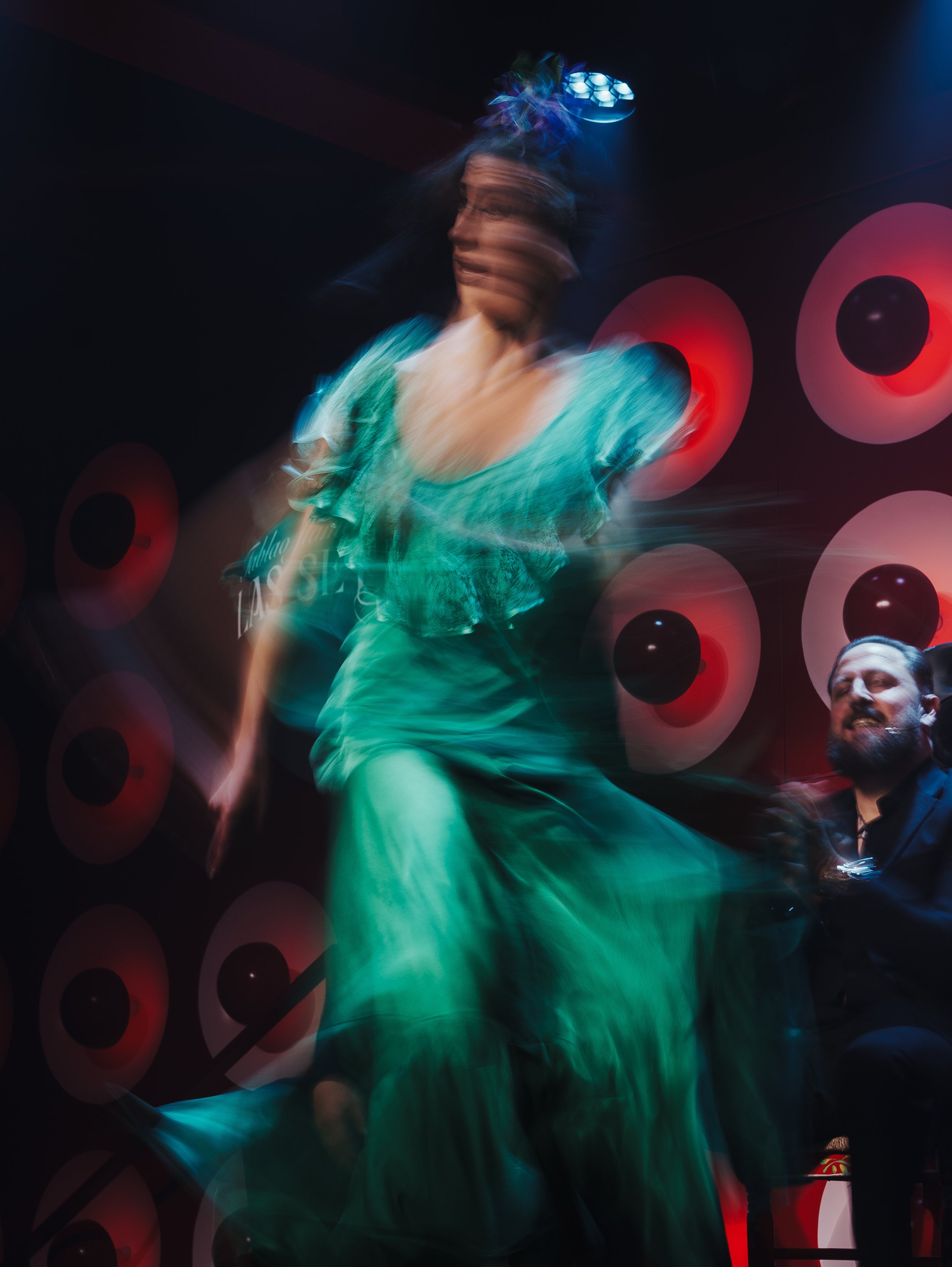 Blurred image of a woman in a turquoise dress dancing at a party or event with circular red and black decorations on the wall in the background.