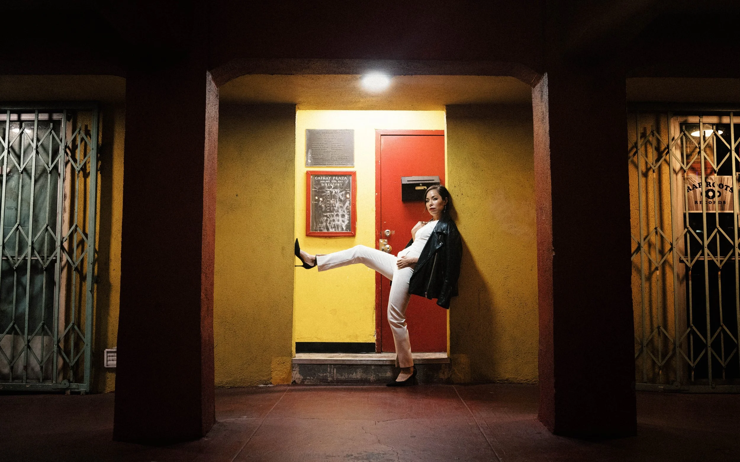 A woman in white pants, a white top, and a black leather jacket, with high heels, is sitting on a red door's threshold in a brightly lit yellow and red building entrance at night, with metal gates on either side.