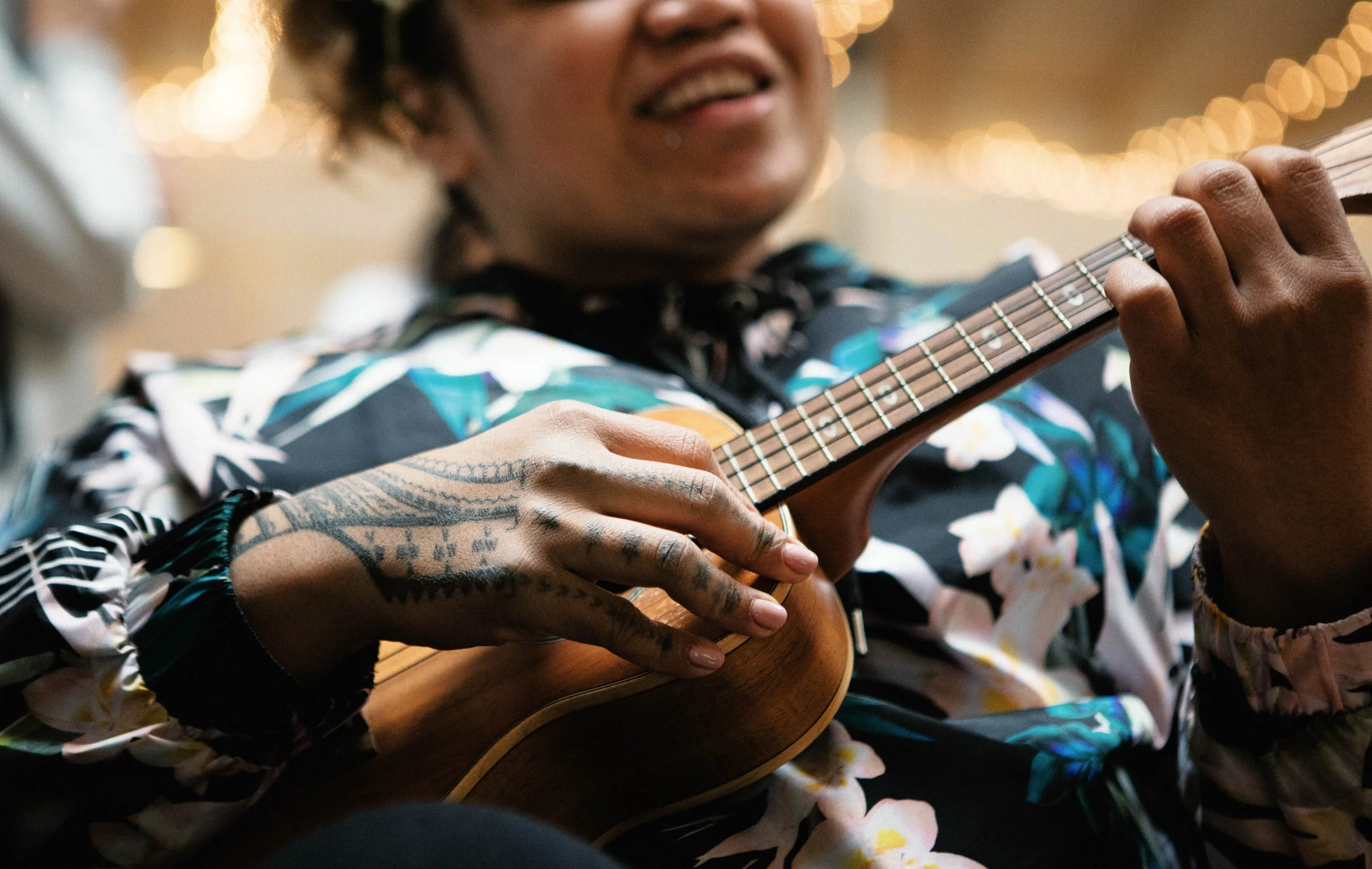 Close-up of a person playing a guitar, wearing a colorful floral shirt, with visible tattoos on their arm. The background features blurred warm string lights.