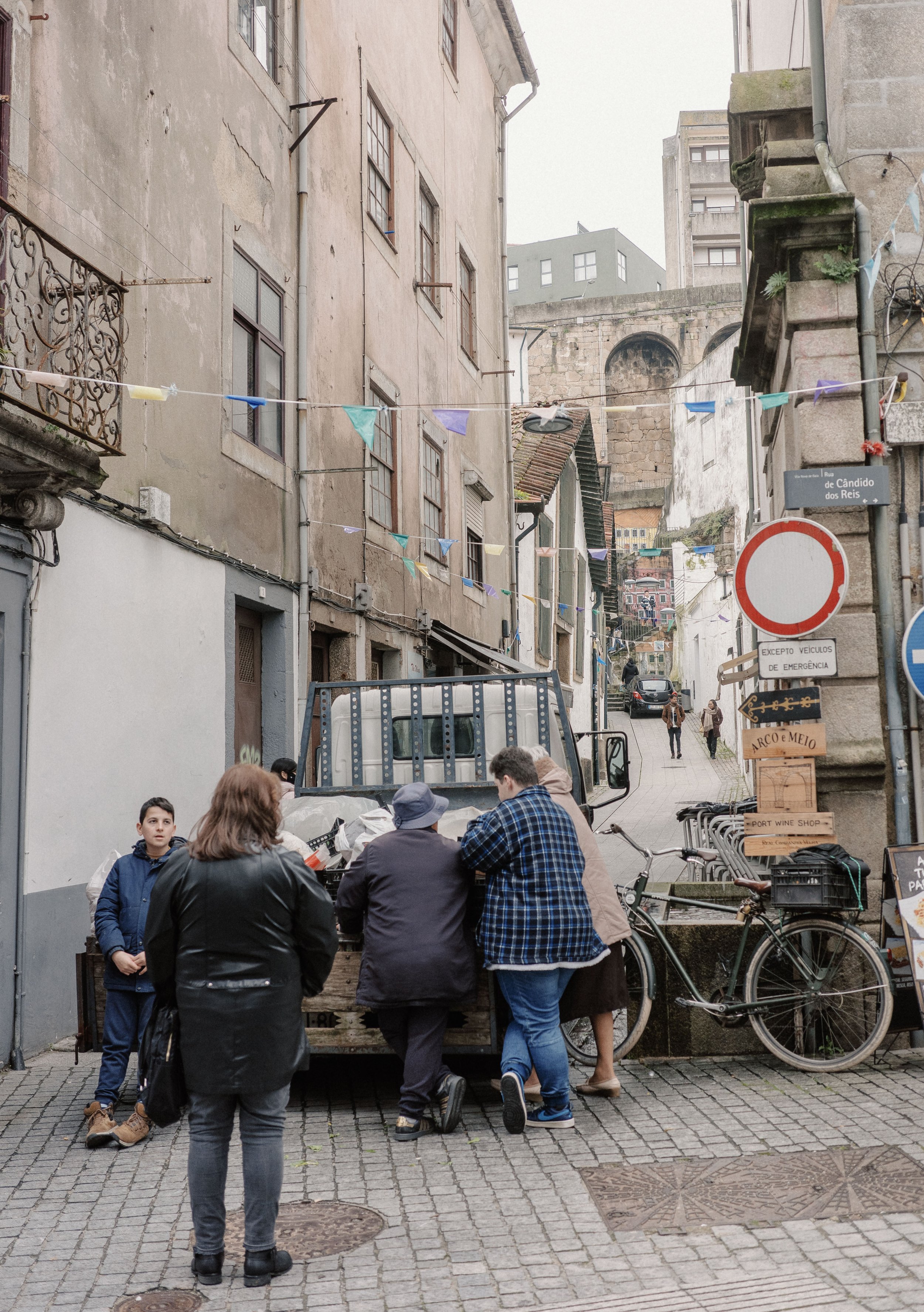 Street scene with people gathered around a small truck, some standing and others sitting, in a narrow, cobblestone alley with old buildings and bicycles nearby, and colorful flags hanging overhead.