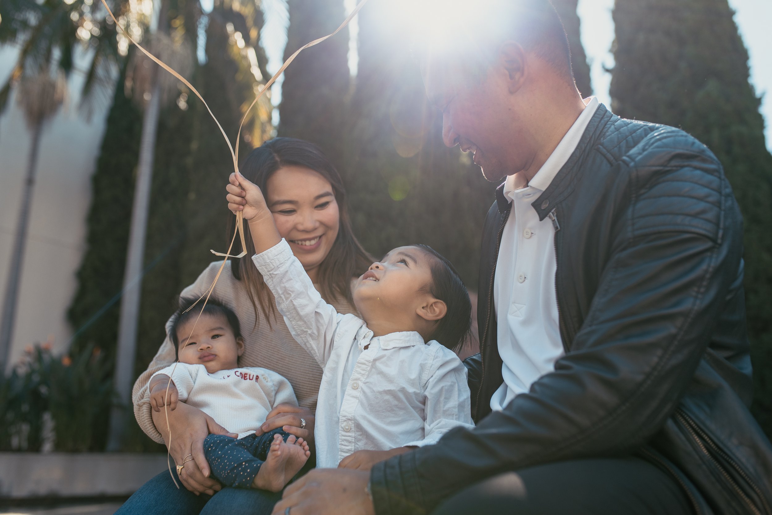 A family of four outdoors with sunlight shining through trees; a man and woman smiling at two young children, one holding a yellow balloon, in a garden or park.