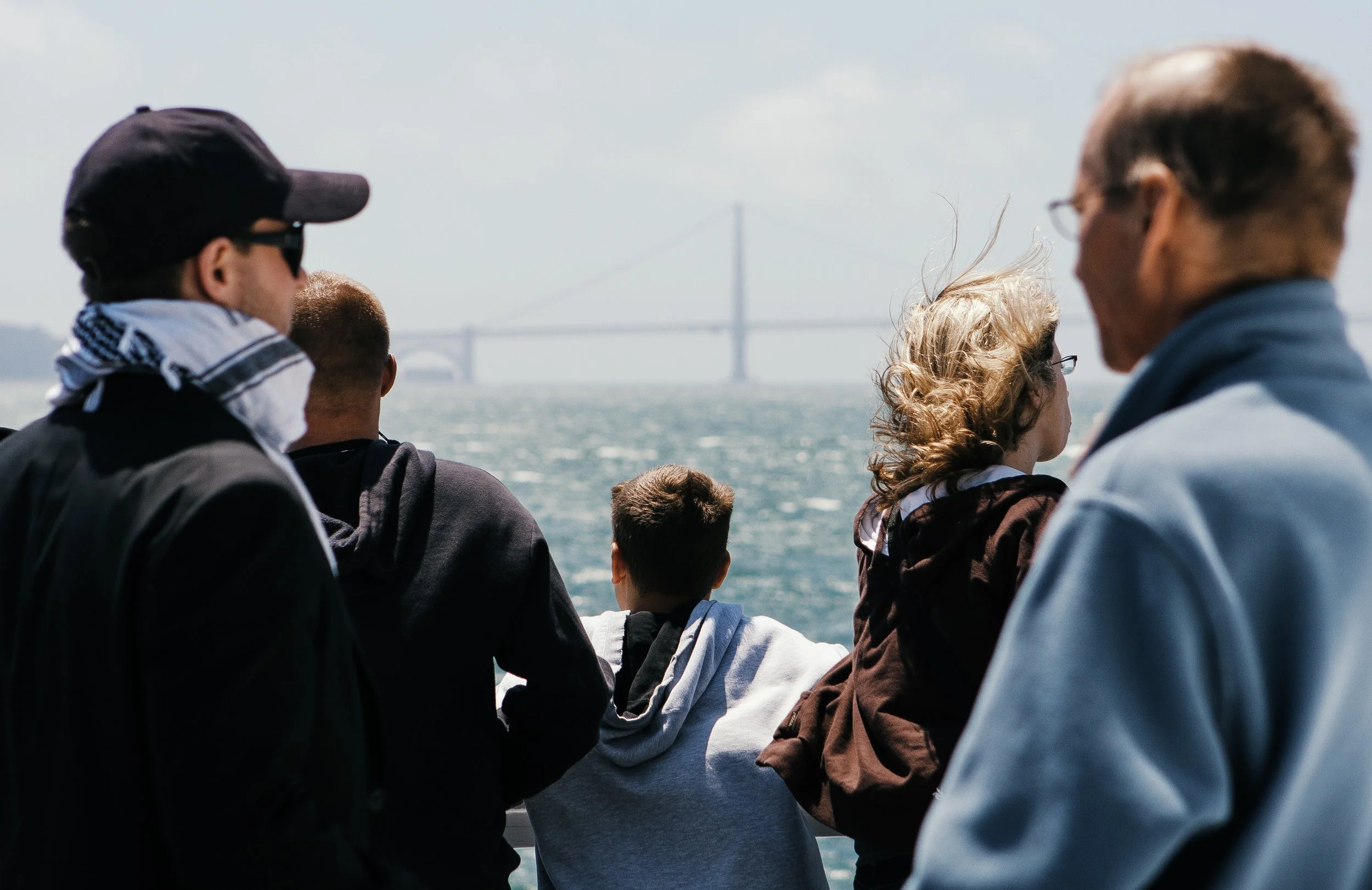 A group of people on a boat or ferry overlooking a large bridge over water in the background.
