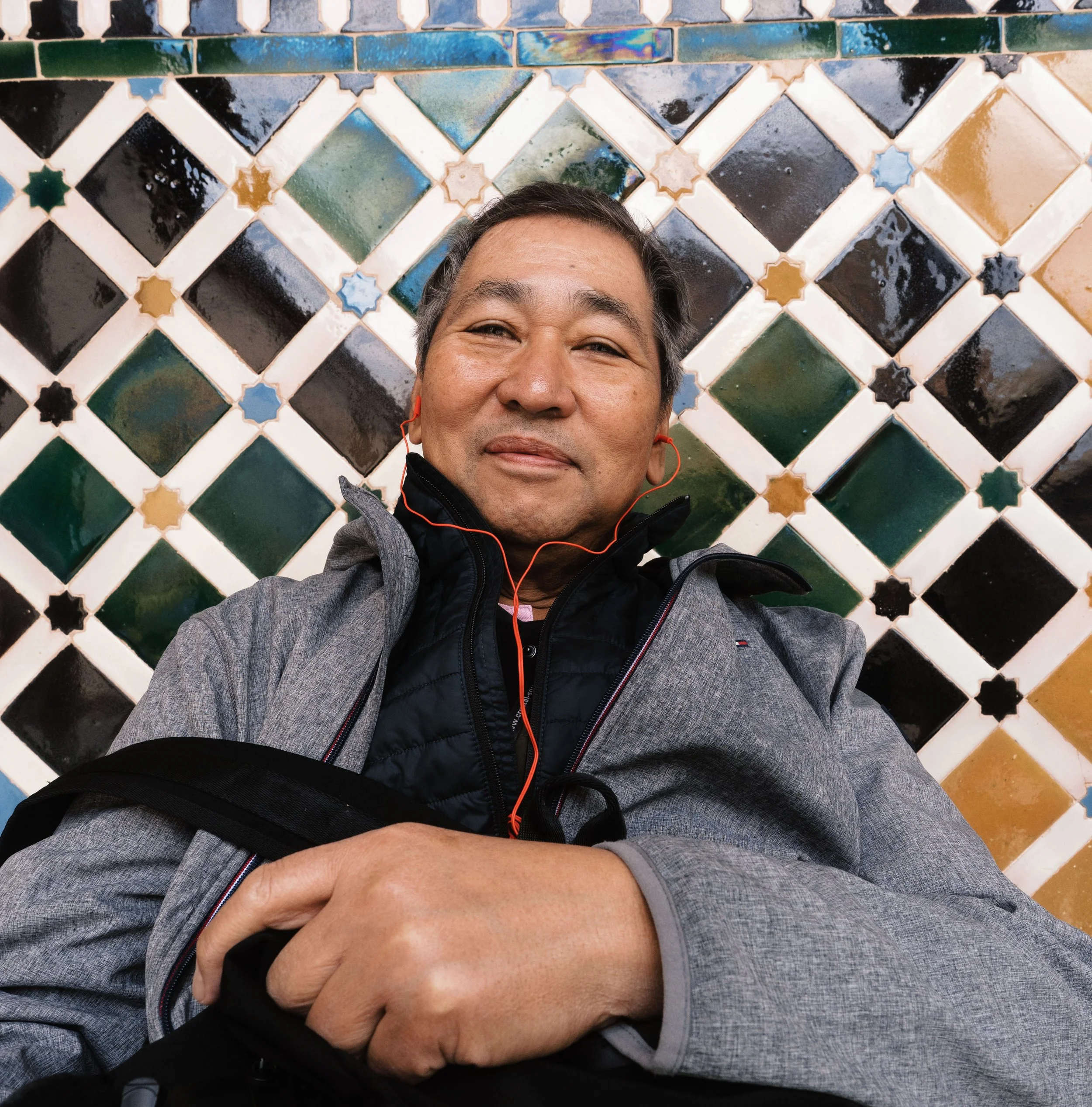 A close-up selfie of a smiling person with short gray hair, wearing gray and black clothing and red earbuds, against a background of colorful glazed tile wall with geometric patterns.