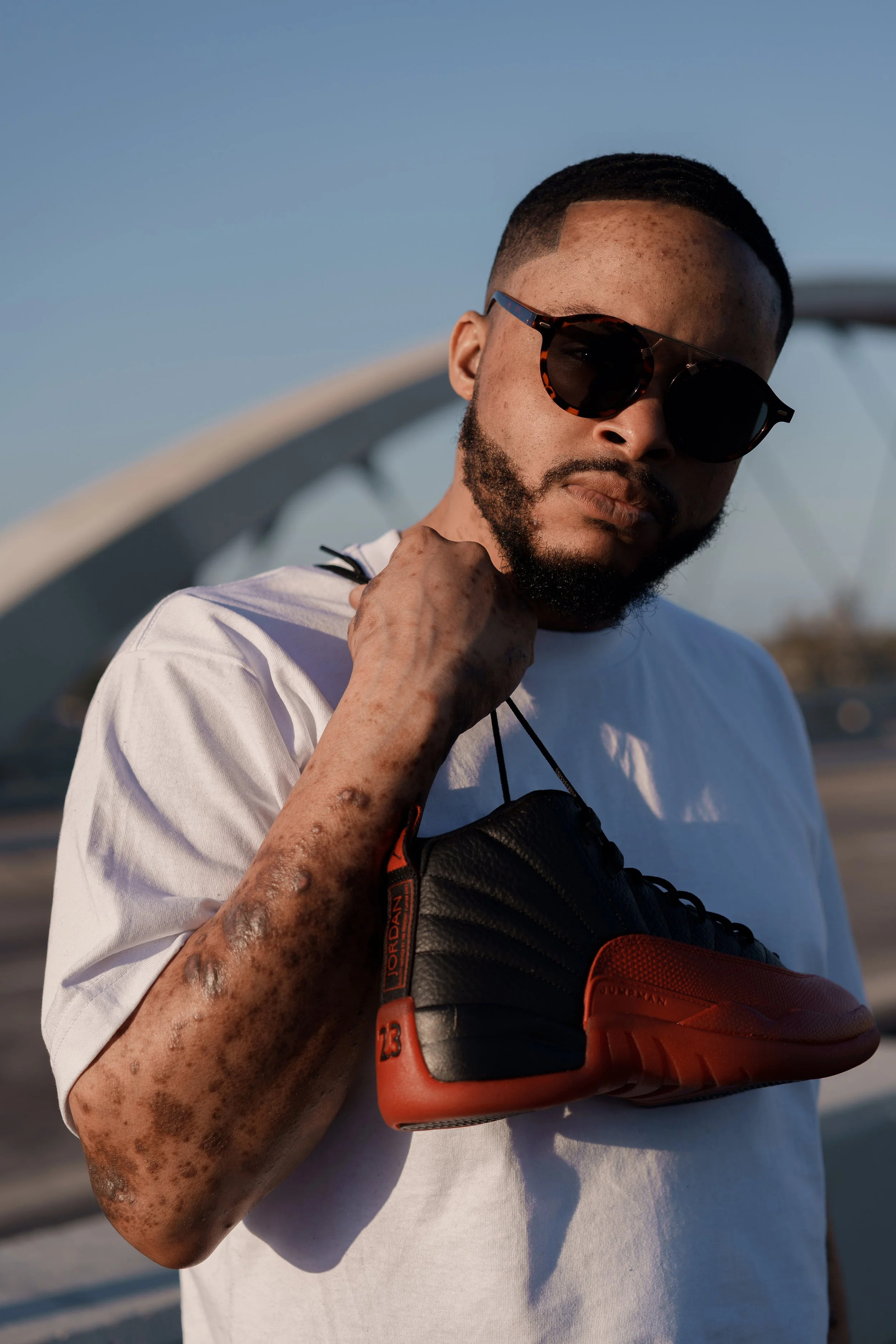A man with short hair and freckles wearing sunglasses, a white t-shirt, and holding a black and red shoe, standing outdoors with a bridge in the background.