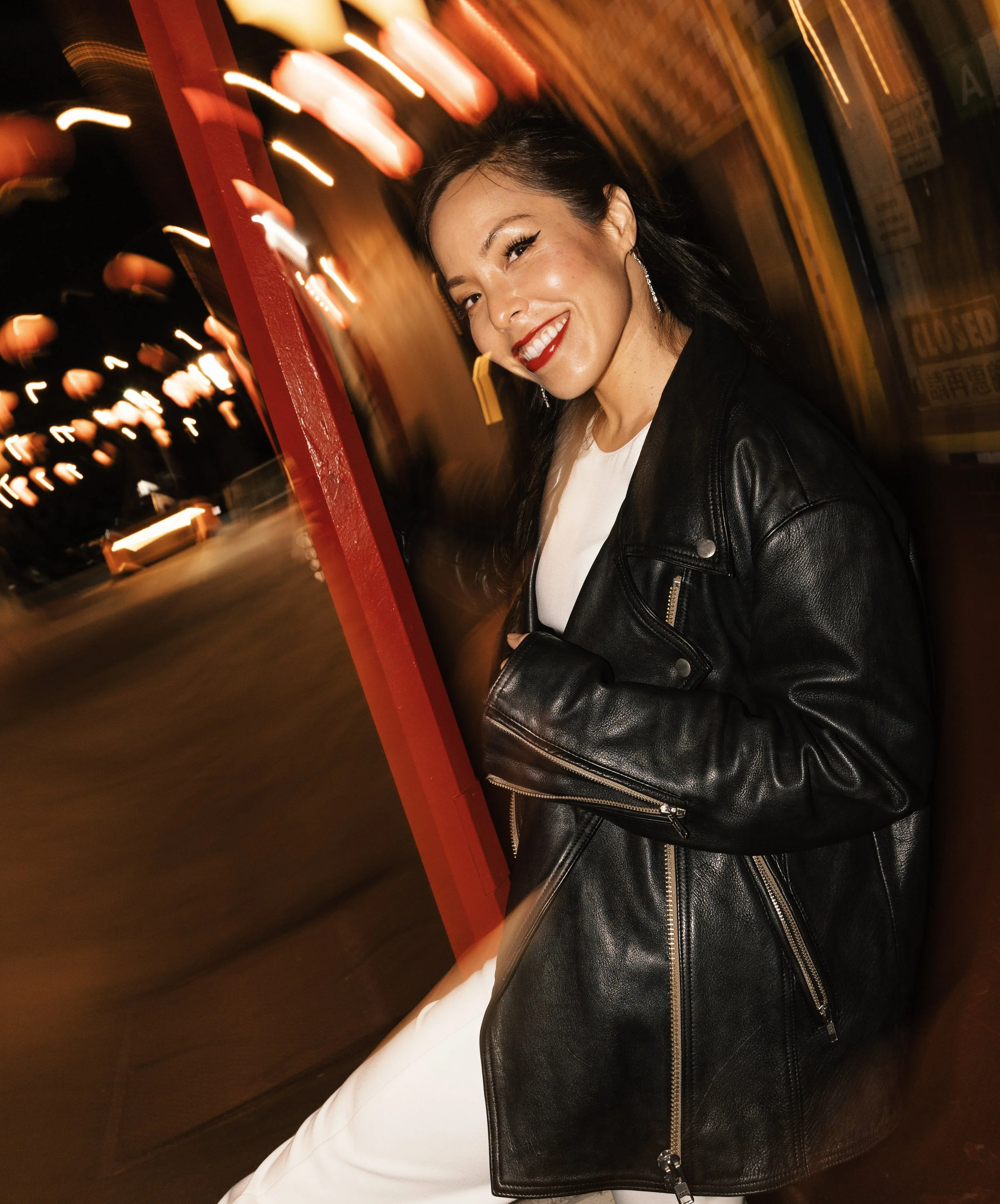 A woman with dark hair, red lipstick, and wearing earrings is smiling in a photo with a blurred, colorful city street background at night. She is wearing a black leather jacket and white pants.