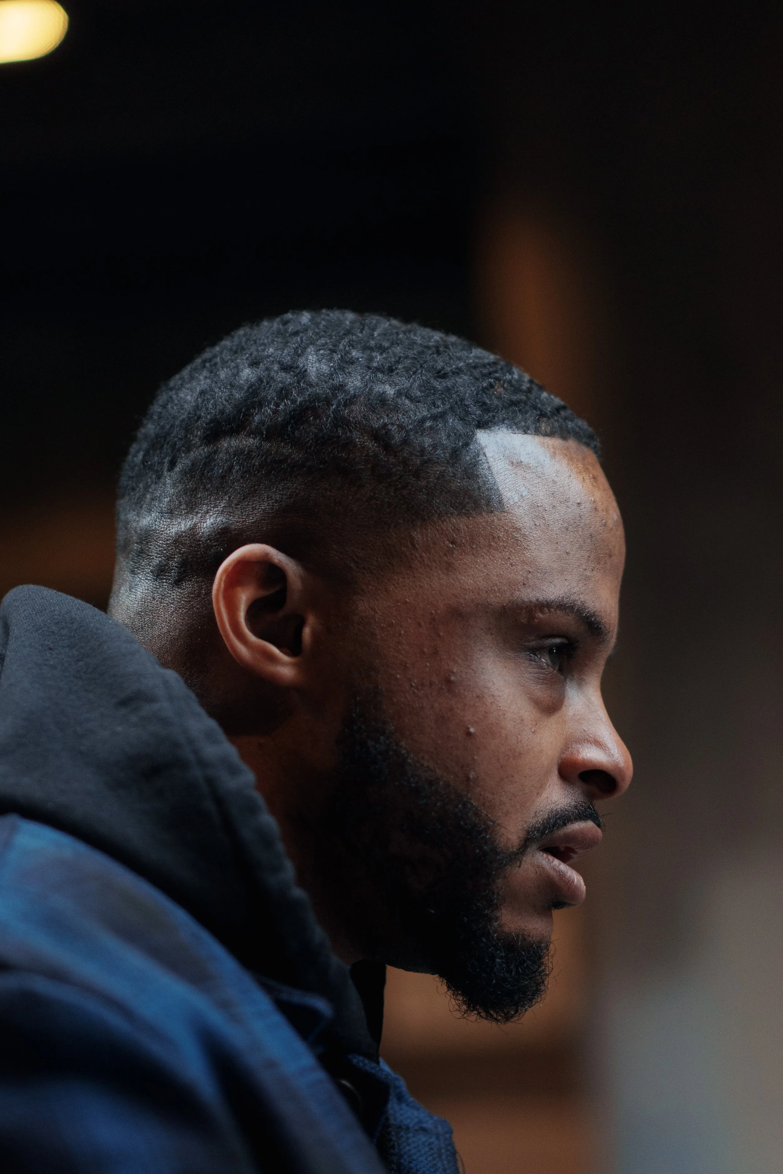 Close-up of a young African-American man with a styled haircut, beard, and mustache, looking to the side against a dark background.