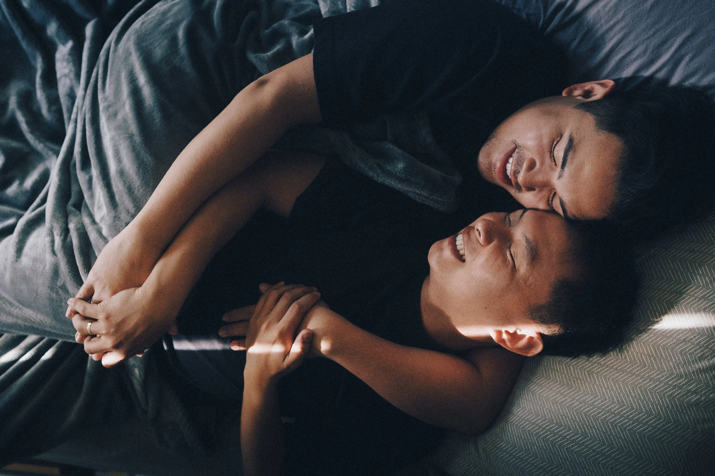 A young man and woman lying on a bed, smiling and holding hands, face to face.