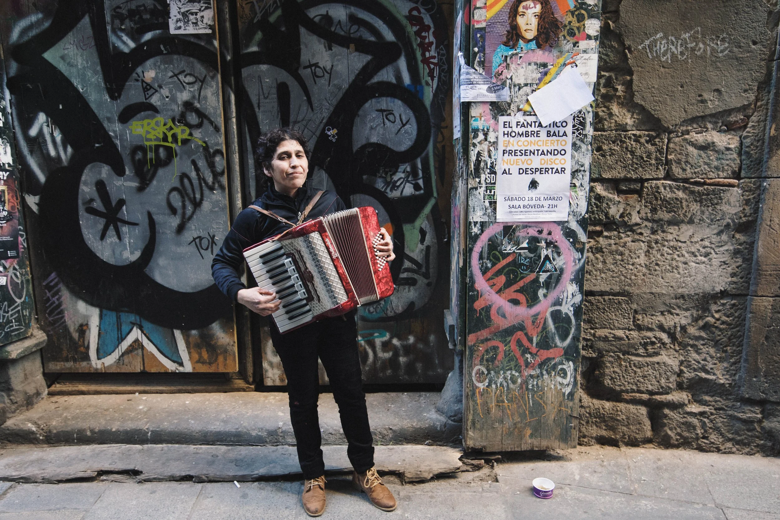 A woman standing in front of a graffiti-covered wall, playing an accordion. She is wearing a black jacket, black pants, and tan boots. There is a paper or flyer posted on the wall to her right, and a small purple container on the ground in front of h